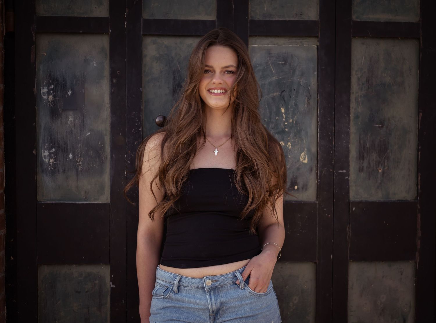 A smiling woman with long wavy brown hair wearing a black crop top and jeans poses against a rustic wooden door.