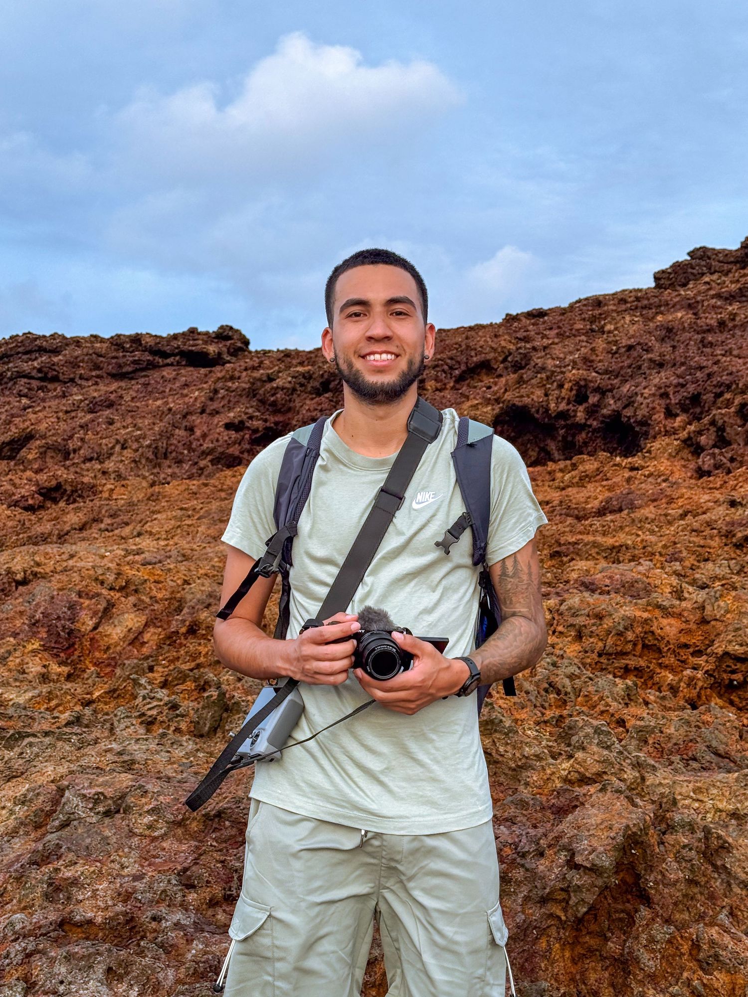 A photographer with camera and backpack stands against reddish-brown rocky terrain under blue sky during a hiking adventure.
