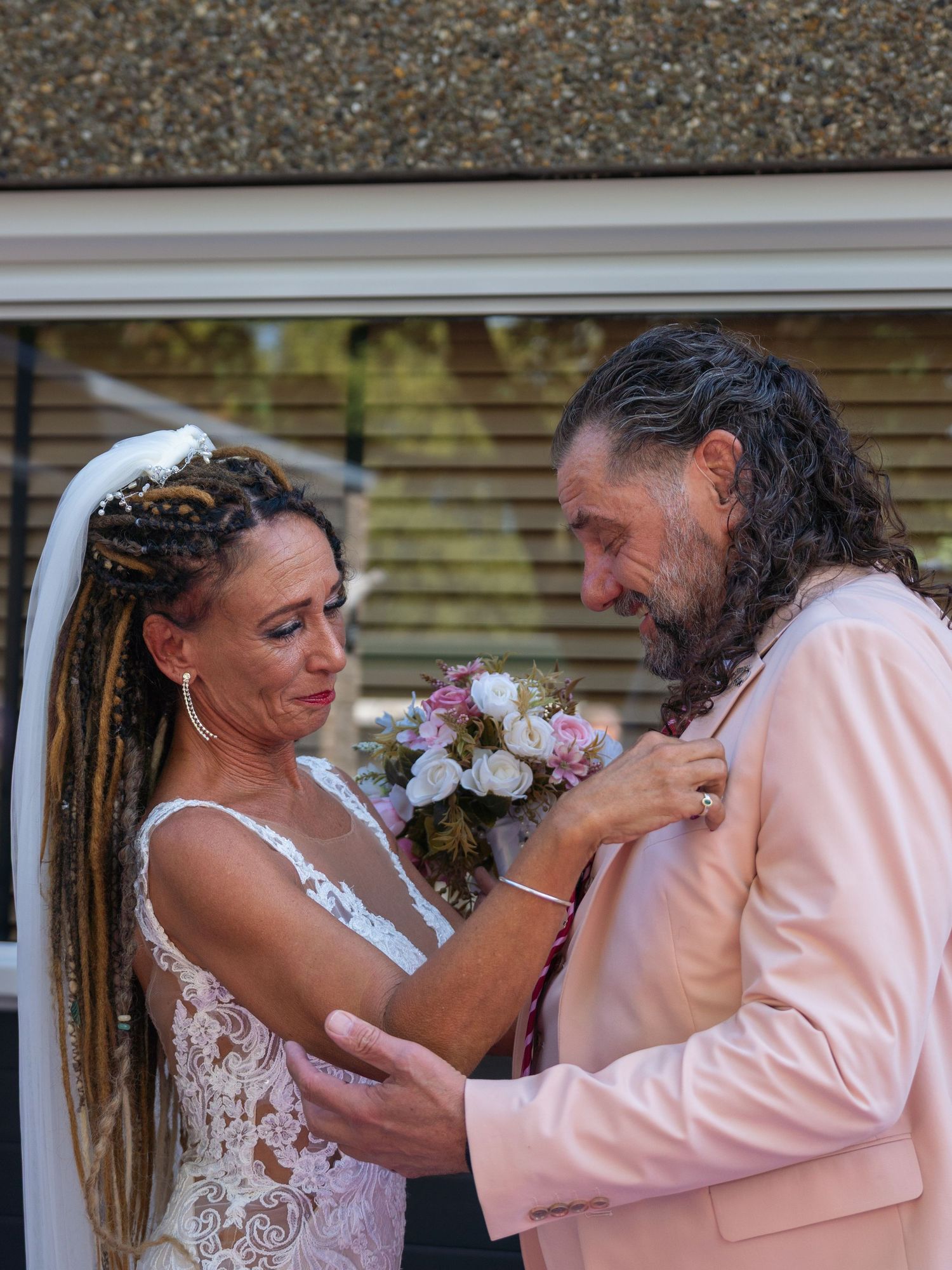 A tender moment between a couple at their wedding, with the bride adjusting the groom's pink suit jacket.