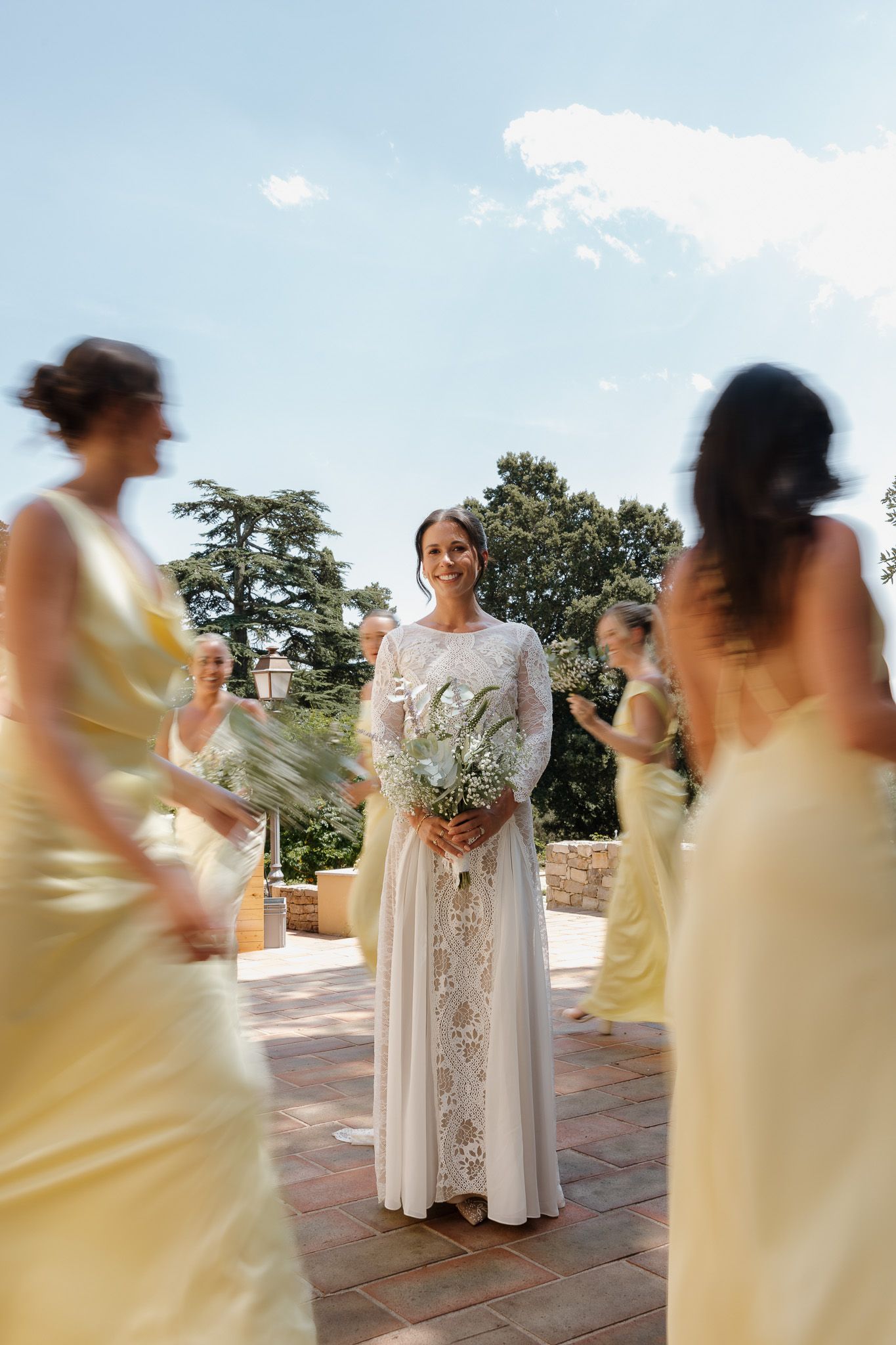 Smiling bride in white lace gown holds bouquet surrounded by bridesmaids in yellow dresses on sunny outdoor patio.