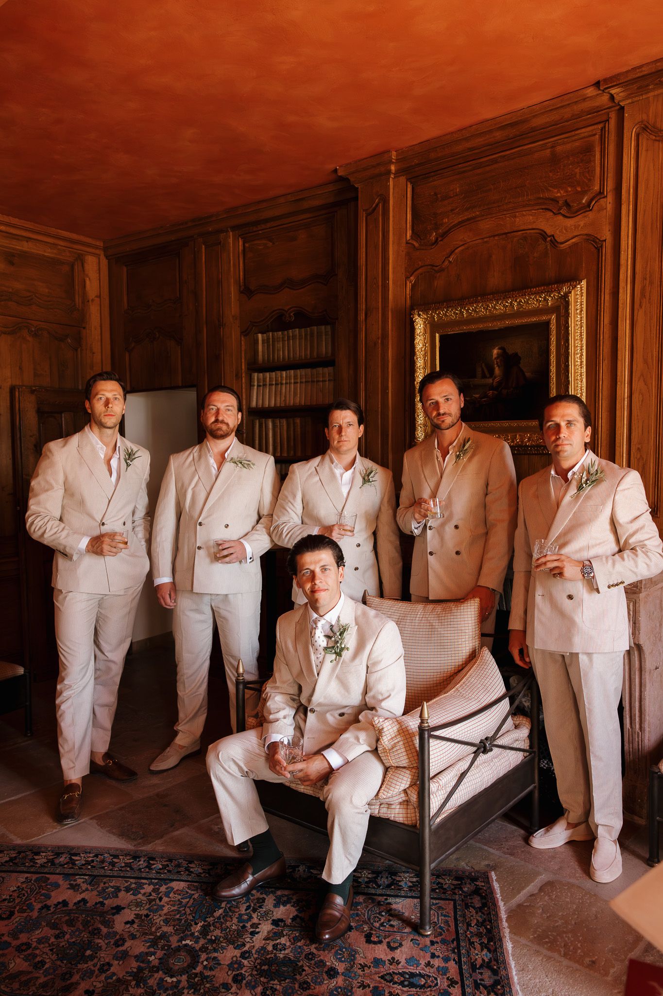 Groom and groomsmen in cream suits with boutonnieres pose in an elegant wood-paneled room with warm lighting.