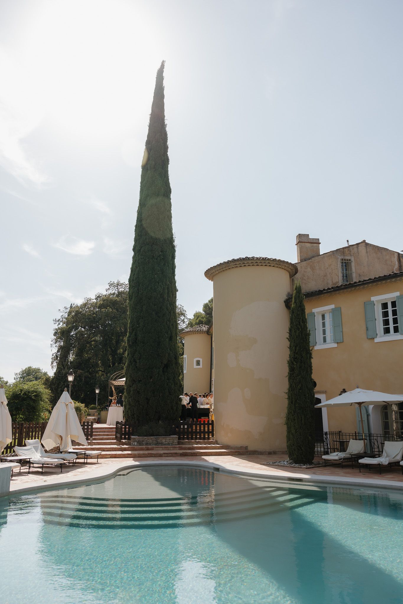 Elegant French chateau with a tall cypress tree beside a sparkling outdoor pool and lounge chairs on a sunny day.