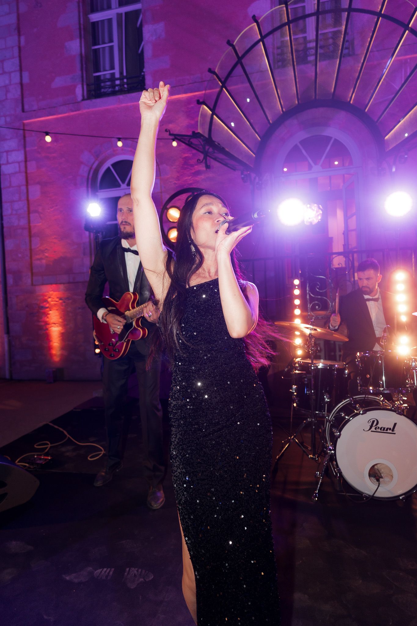 Female singer in black sequin gown performing on stage with hand raised, guitarist and drummer visible behind her.