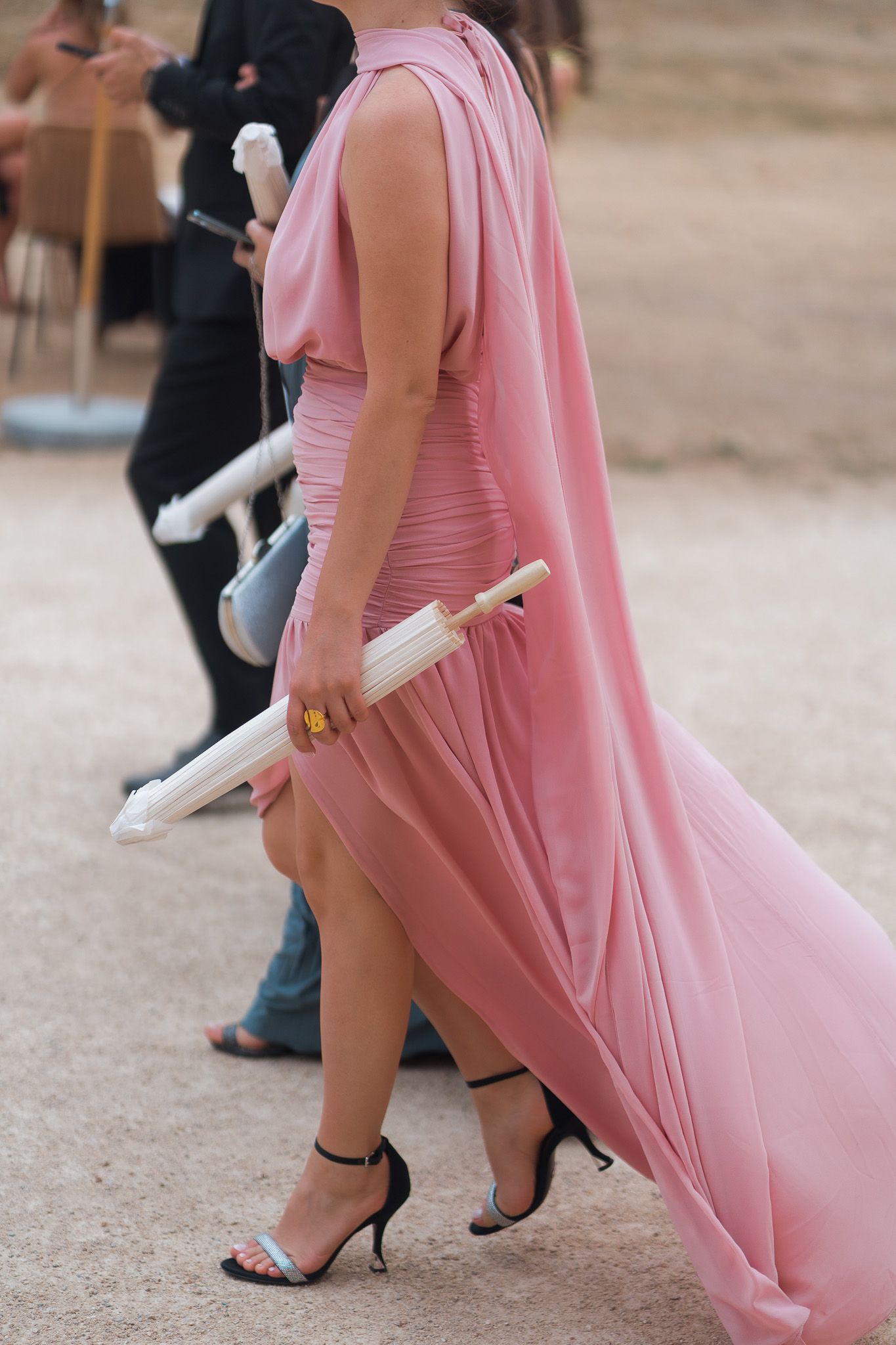Woman in elegant pink flowing gown and black heels holding a white parasol at an outdoor event.