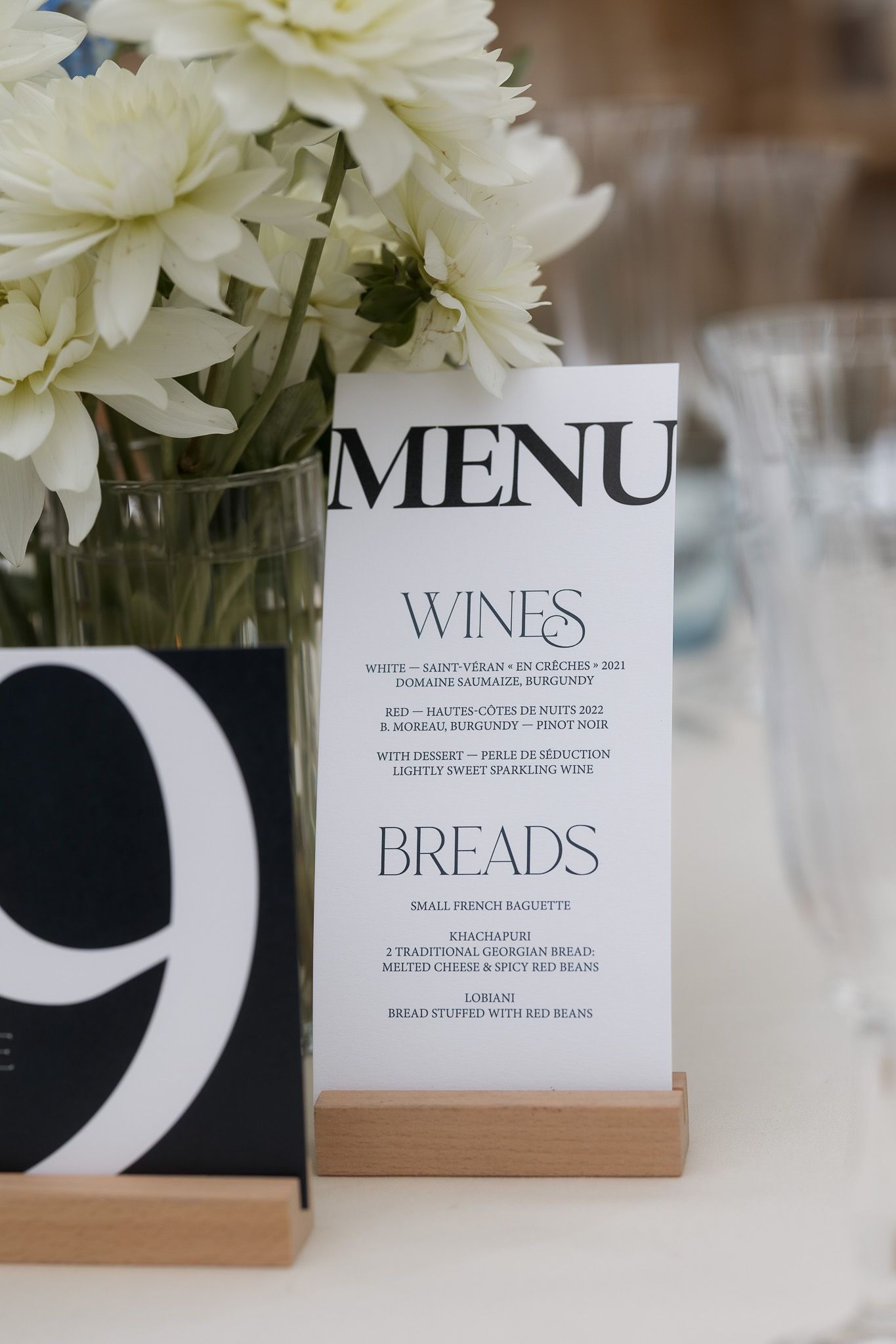 Elegant wedding menu card in wooden holder listing wines and breads beside white floral centerpiece at table nine.