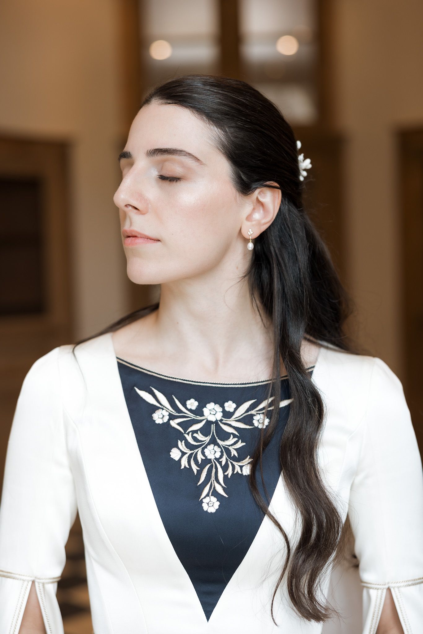 Elegant woman in white jacket with dark floral-embroidered panel, pearl earring, hair accessory, eyes closed.