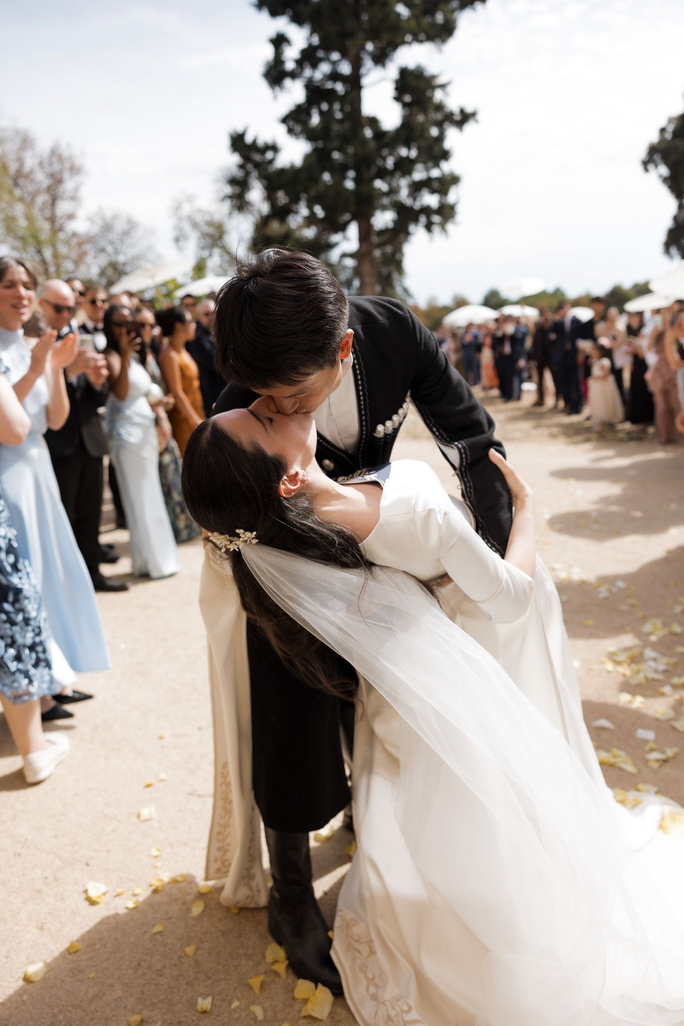 Groom dips bride for a kiss at outdoor wedding ceremony while guests applaud in background.