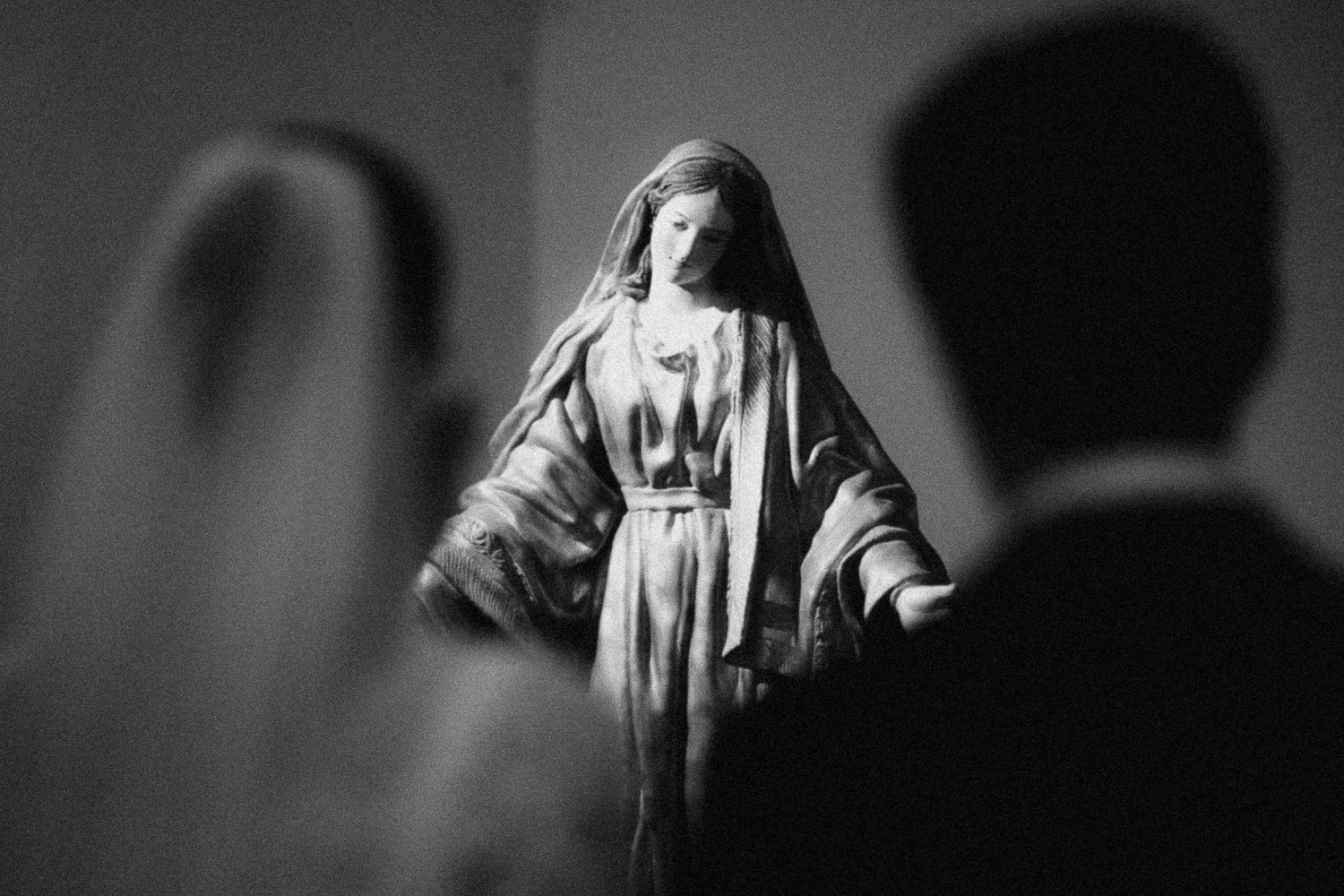 Black and white photo of a Virgin Mary statue with blurred wedding couple in foreground at a church ceremony.