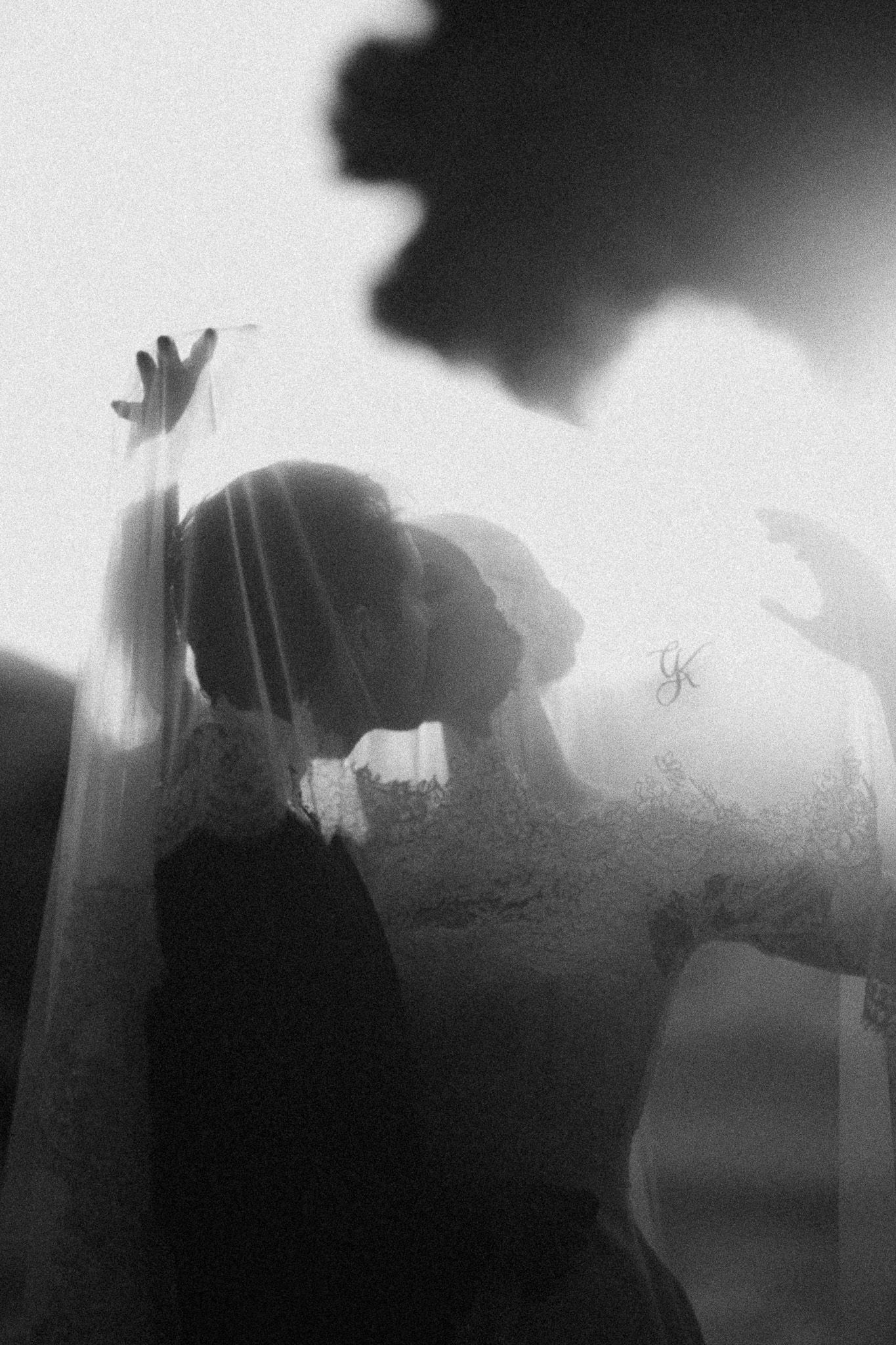 Artistic black and white wedding photo of bride kissing under a flowing veil.