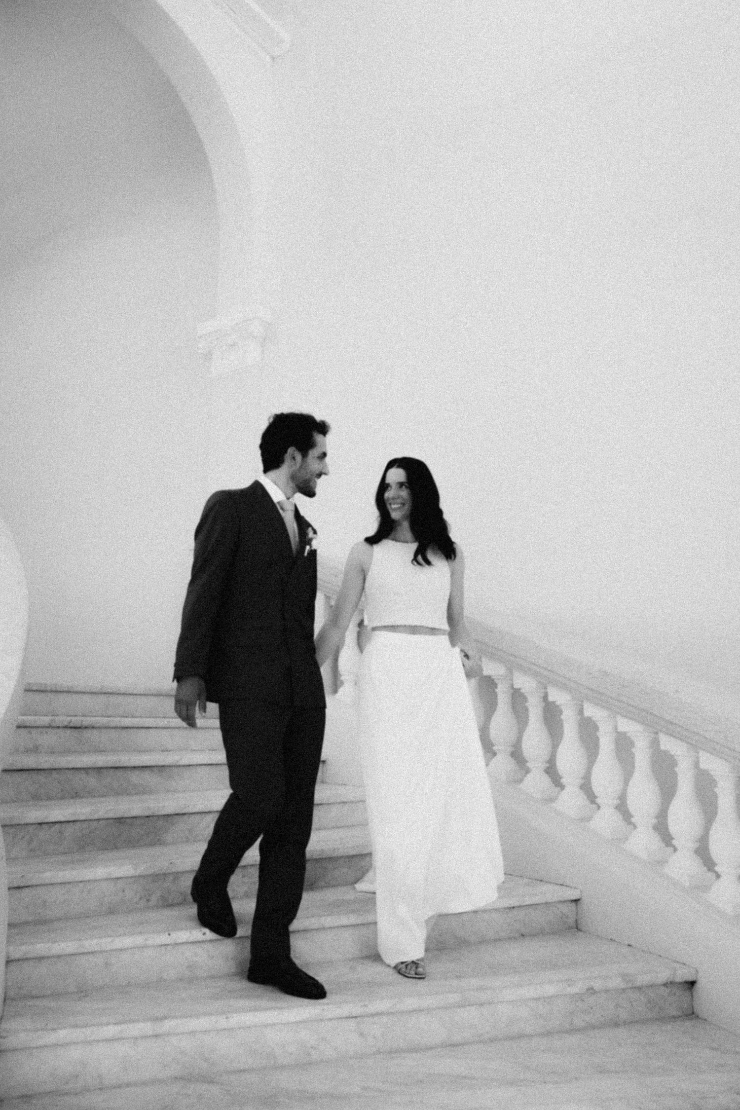 Black and white photo of a bride and groom holding hands on elegant marble stairs at their wedding.