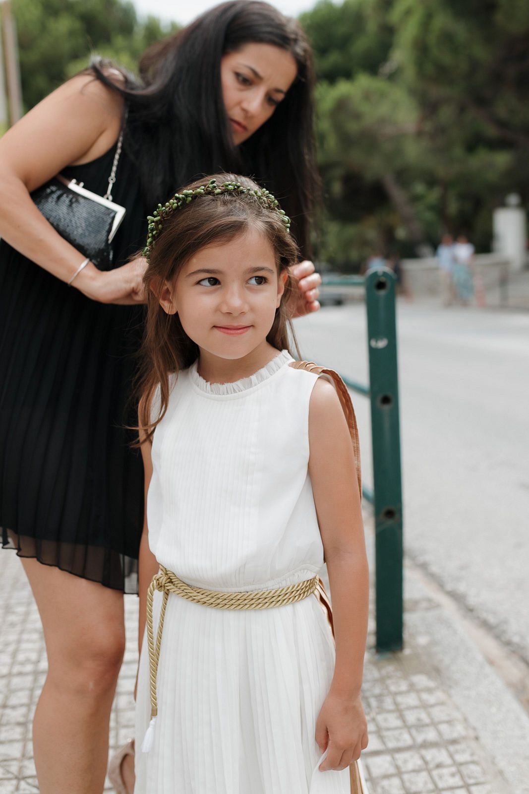 A young flower girl in a white dress with a gold belt stands on a stone path outdoors.