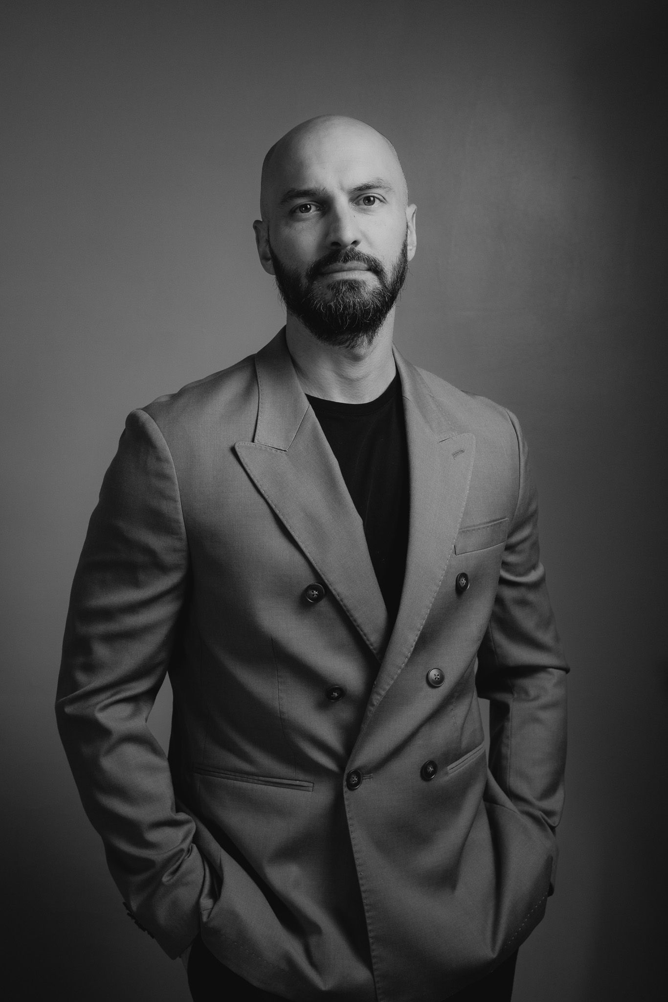 Black and white studio portrait of a bald, bearded man in a double-breasted blazer and dark t-shirt, hands in pockets.
