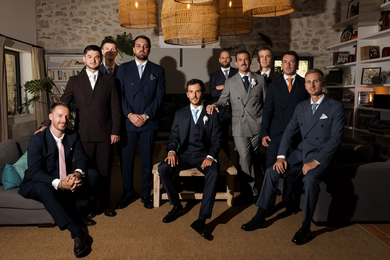 Group of nine groomsmen posing together in stylish suits indoors for a wedding portrait.