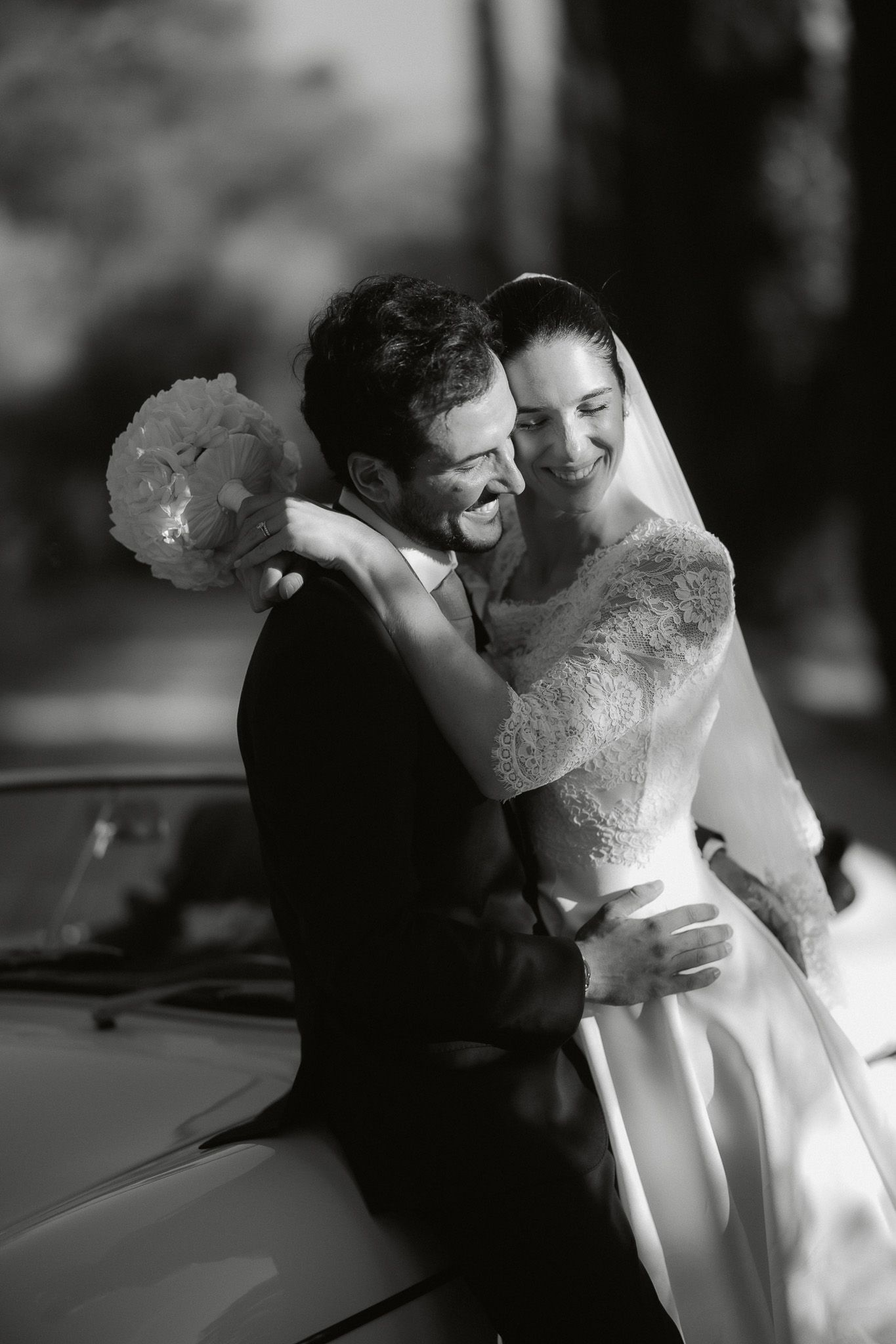 Black and white photo of a smiling bride and groom embracing near a vintage car on their wedding day.