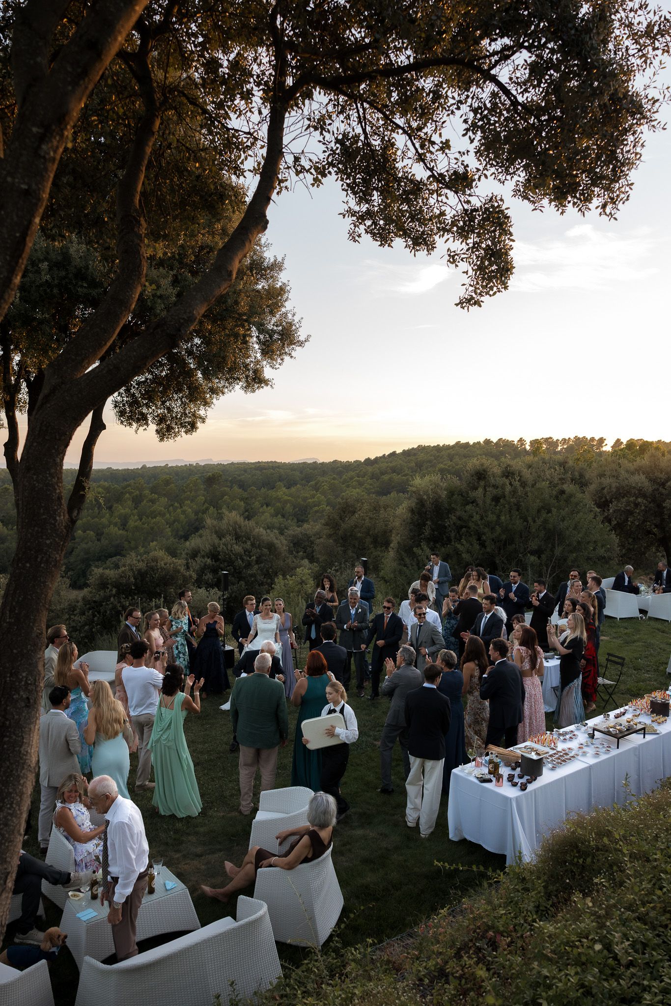 Outdoor wedding cocktail hour at sunset with elegantly dressed guests mingling on a terrace overlooking lush green landscape.