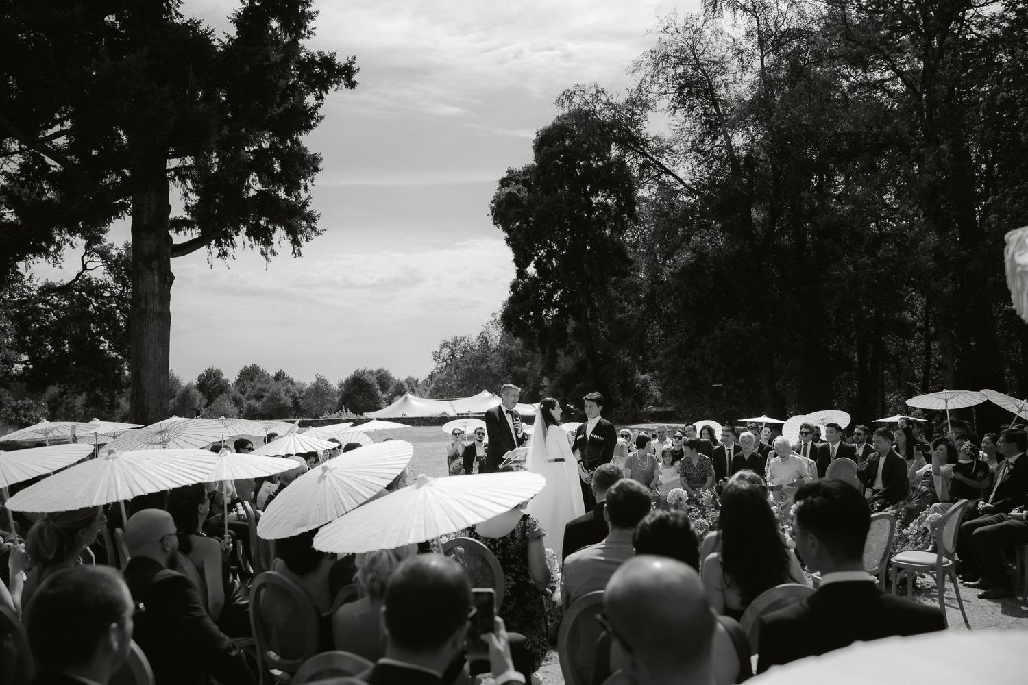 Black and white outdoor wedding ceremony with guests seated under white parasols surrounded by lush trees.