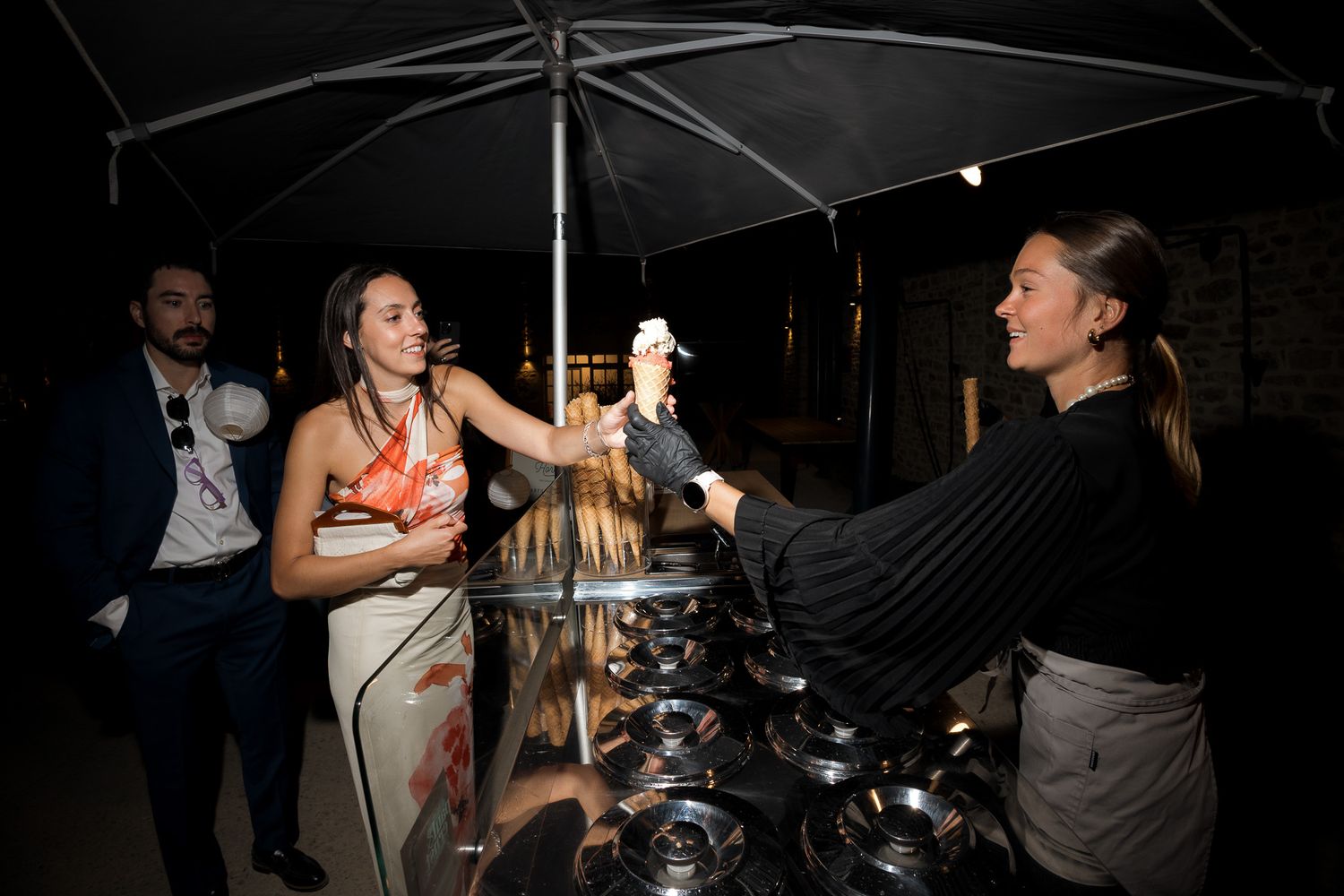 Woman receiving ice cream from server at outdoor night event under umbrella, with man in suit watching nearby.