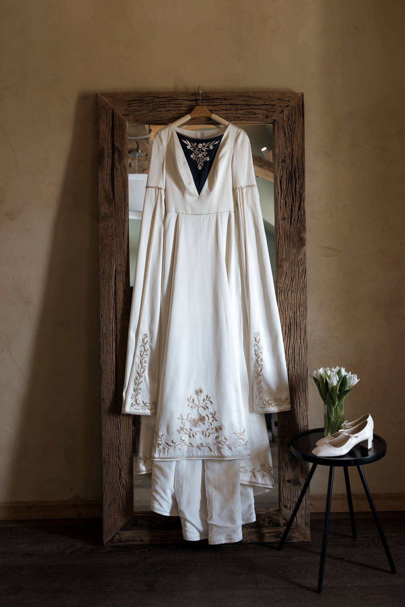 White wedding dress with floral embroidery hanging on rustic mirror frame, with bridal heels and tulips on black side table.