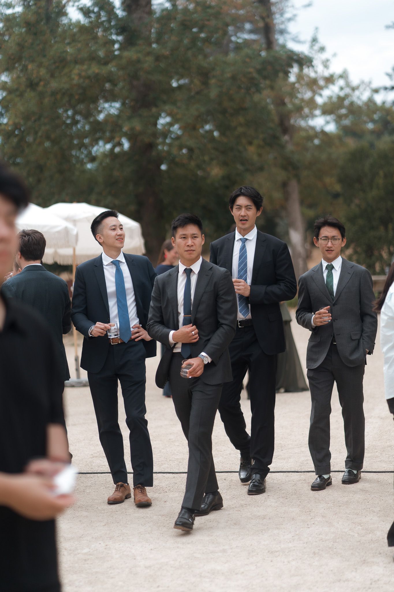 Four Asian men in dark suits and blue ties walking outdoors at a formal event, trees visible in background.