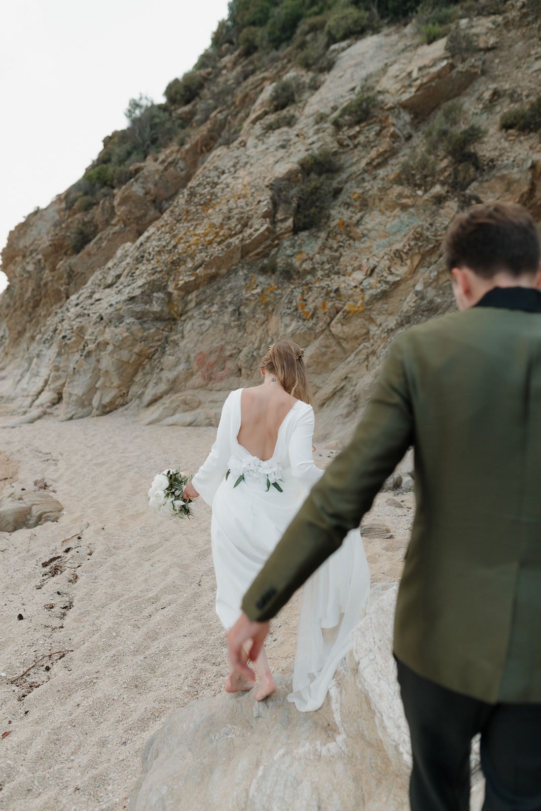 A couple in wedding attire walks along a sandy beach path next to rocky cliffs on their special day.