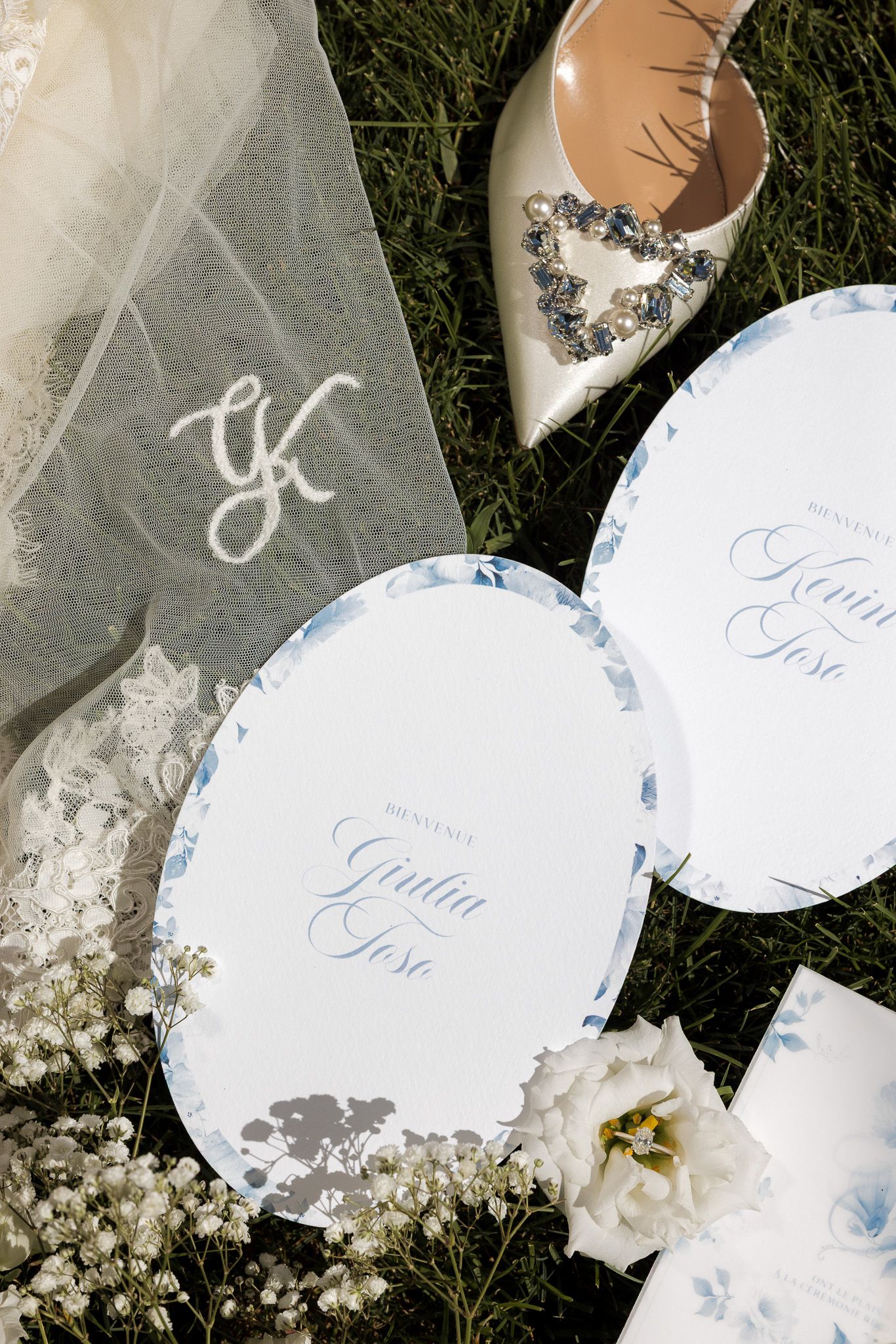 Elegant wedding flat lay with monogram veil, jeweled heels, blue floral place cards reading Giulia Jose on green grass.