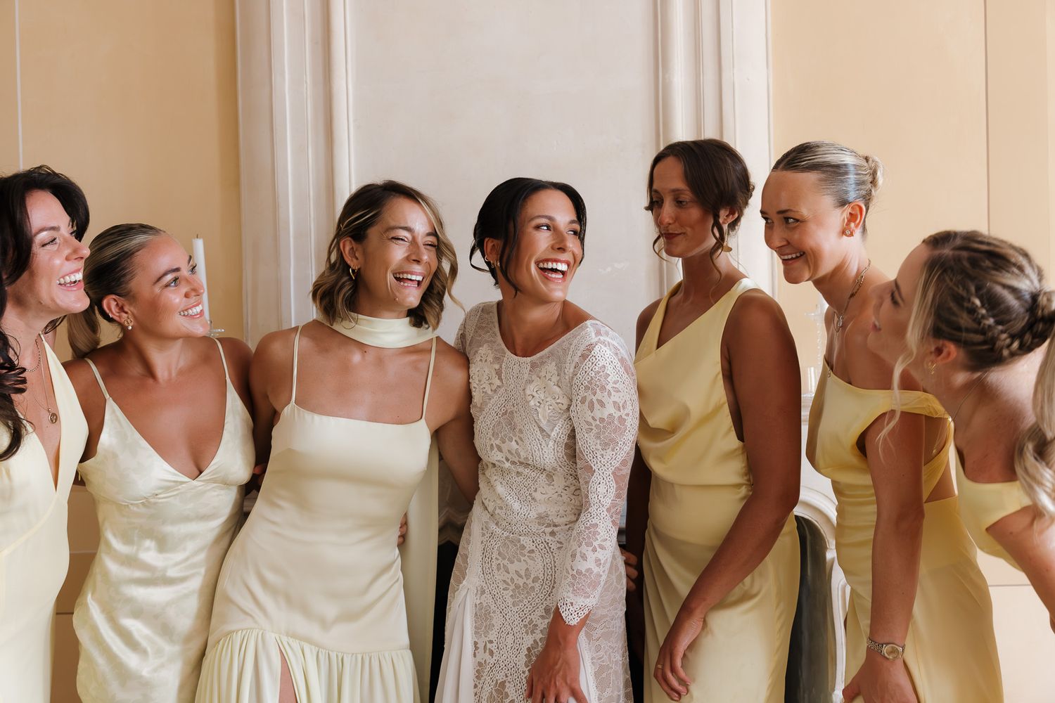 Bride in lace gown laughing with bridesmaids in yellow satin dresses at elegant wedding celebration.