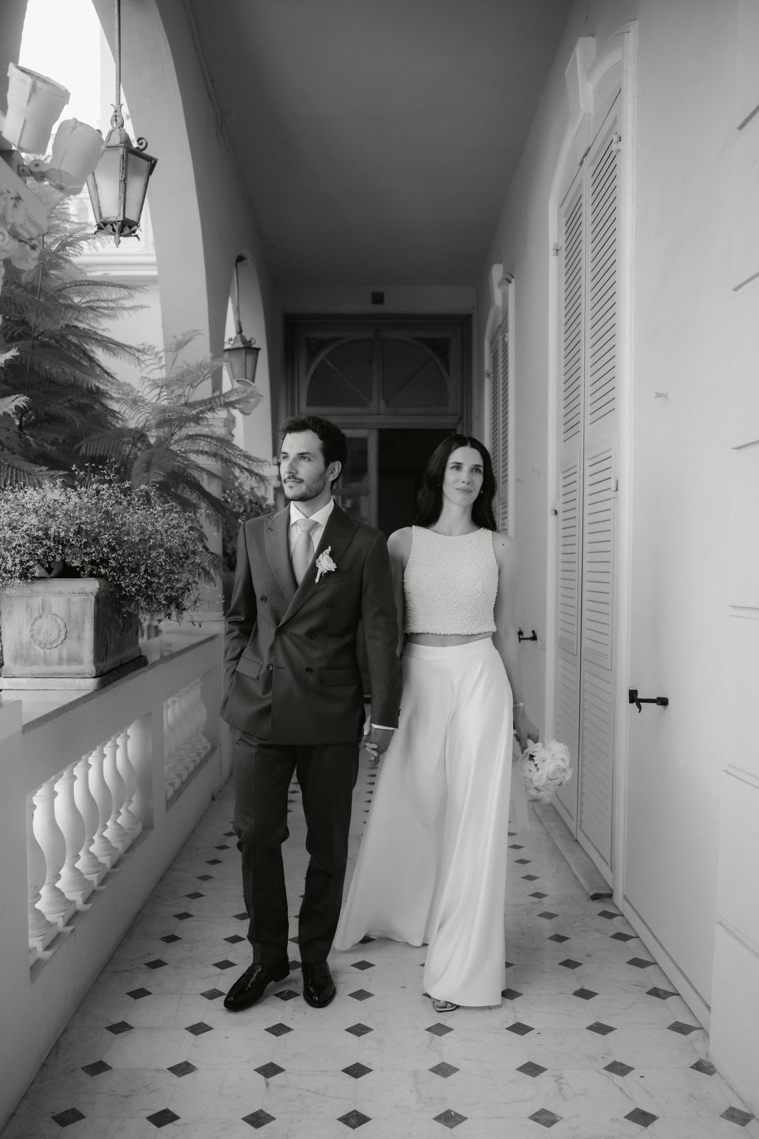 Black and white photo of bride and groom holding hands on elegant tiled veranda with lush plants.