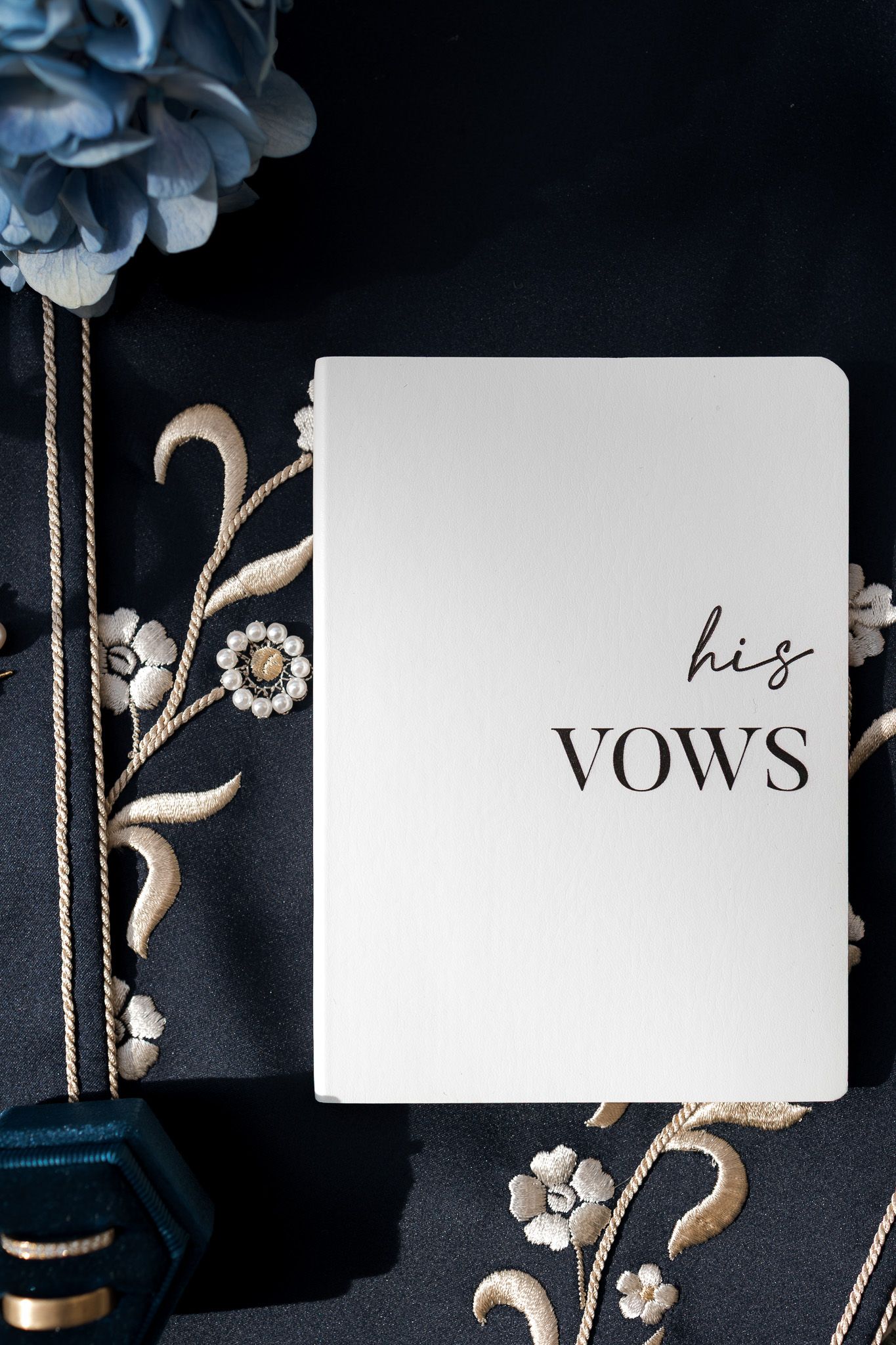 White wedding vow booklet labeled his vows on dark floral embroidered fabric with blue hydrangeas.