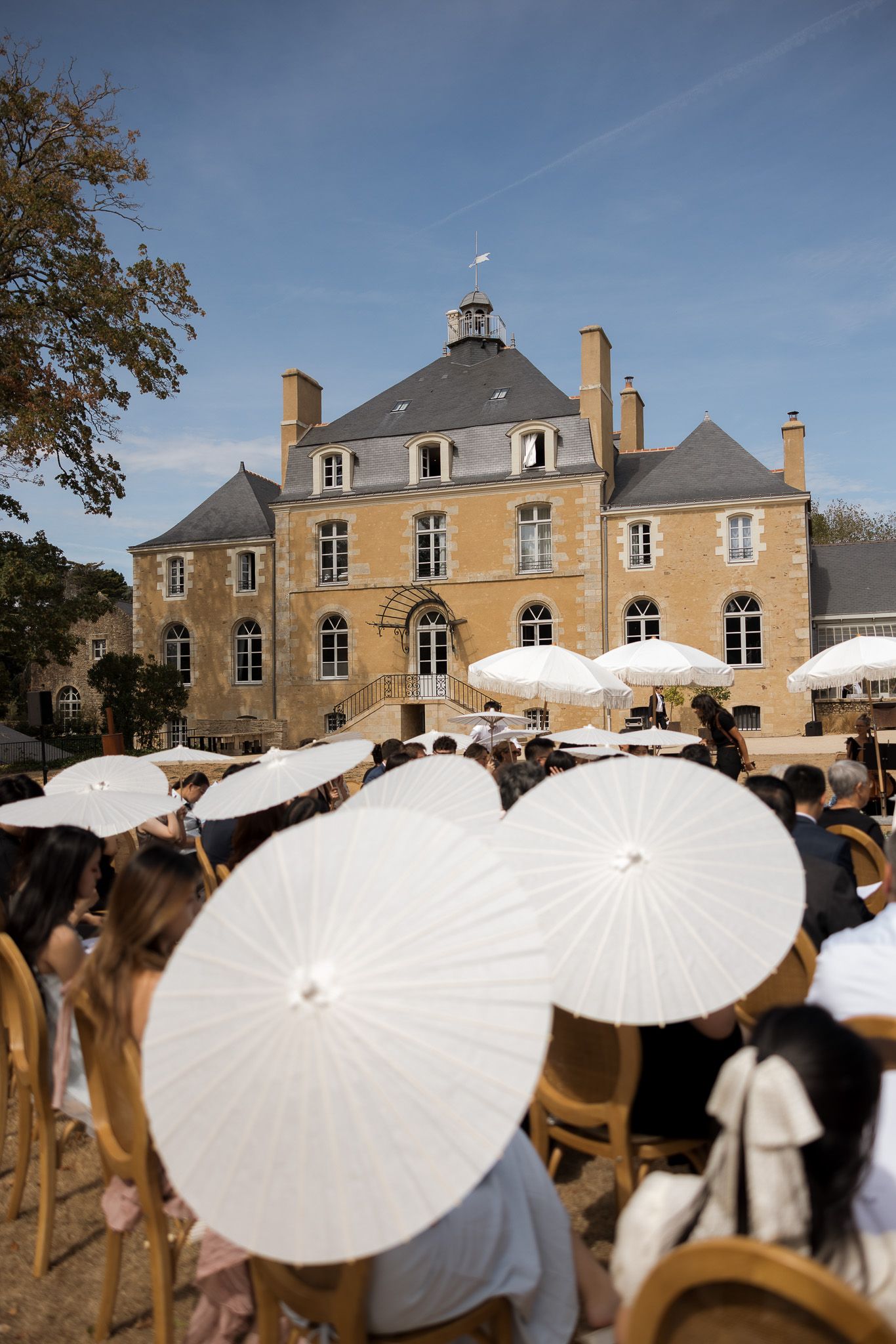 Outdoor wedding ceremony at a French chateau with guests holding white parasols under a blue sky.