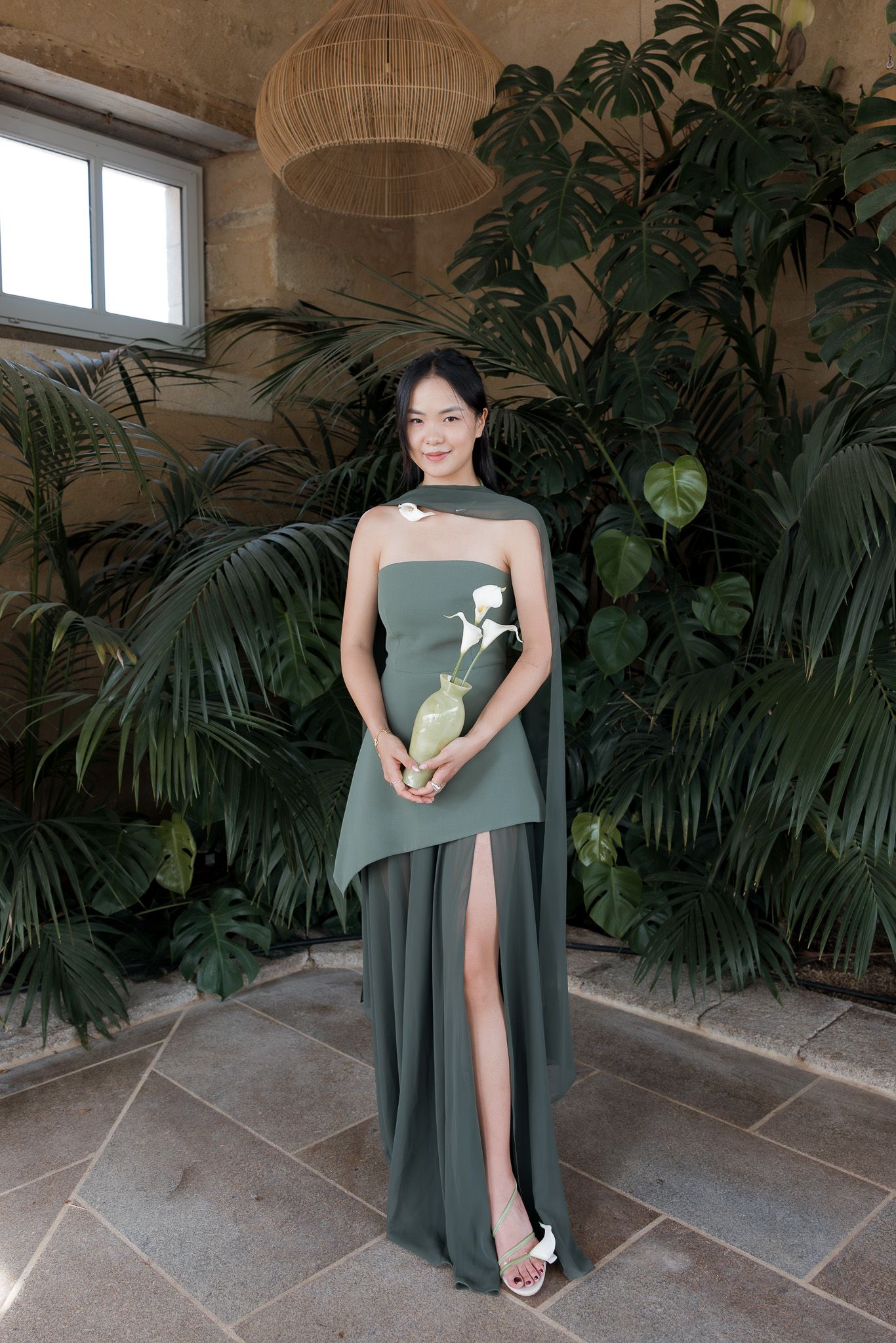 Woman in elegant green asymmetric gown holding a vase with white flowers, posing before lush tropical plants.