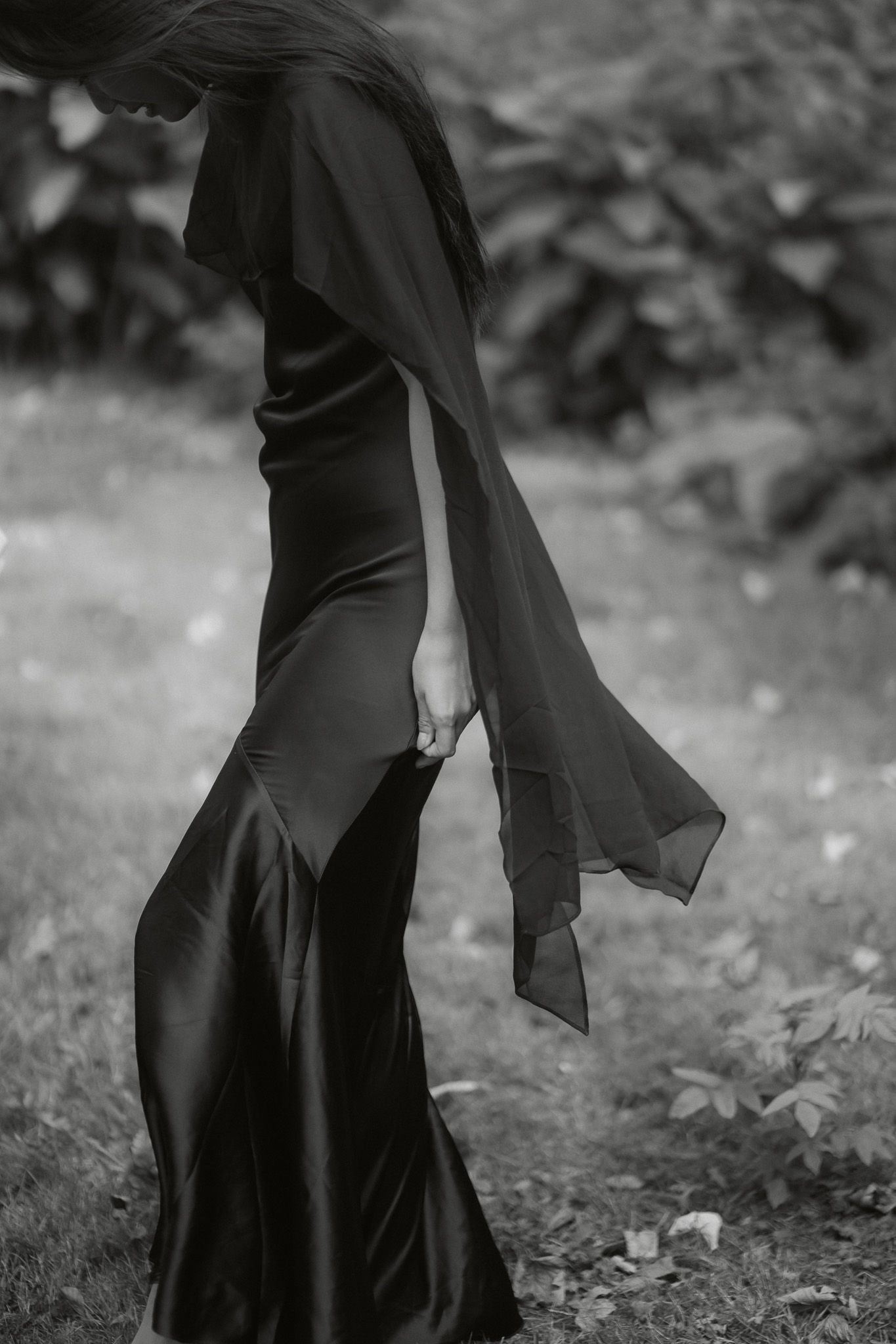 Elegant woman in flowing black satin gown with dramatic cape sleeves poses in garden, black and white photo.