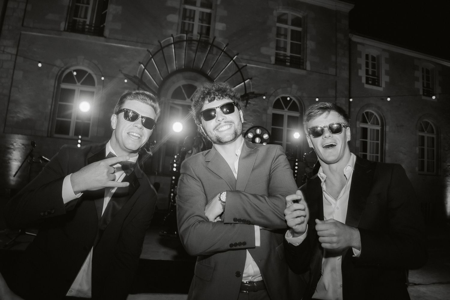 Three men in suits and sunglasses pose playfully outdoors at night in a black and white photo.