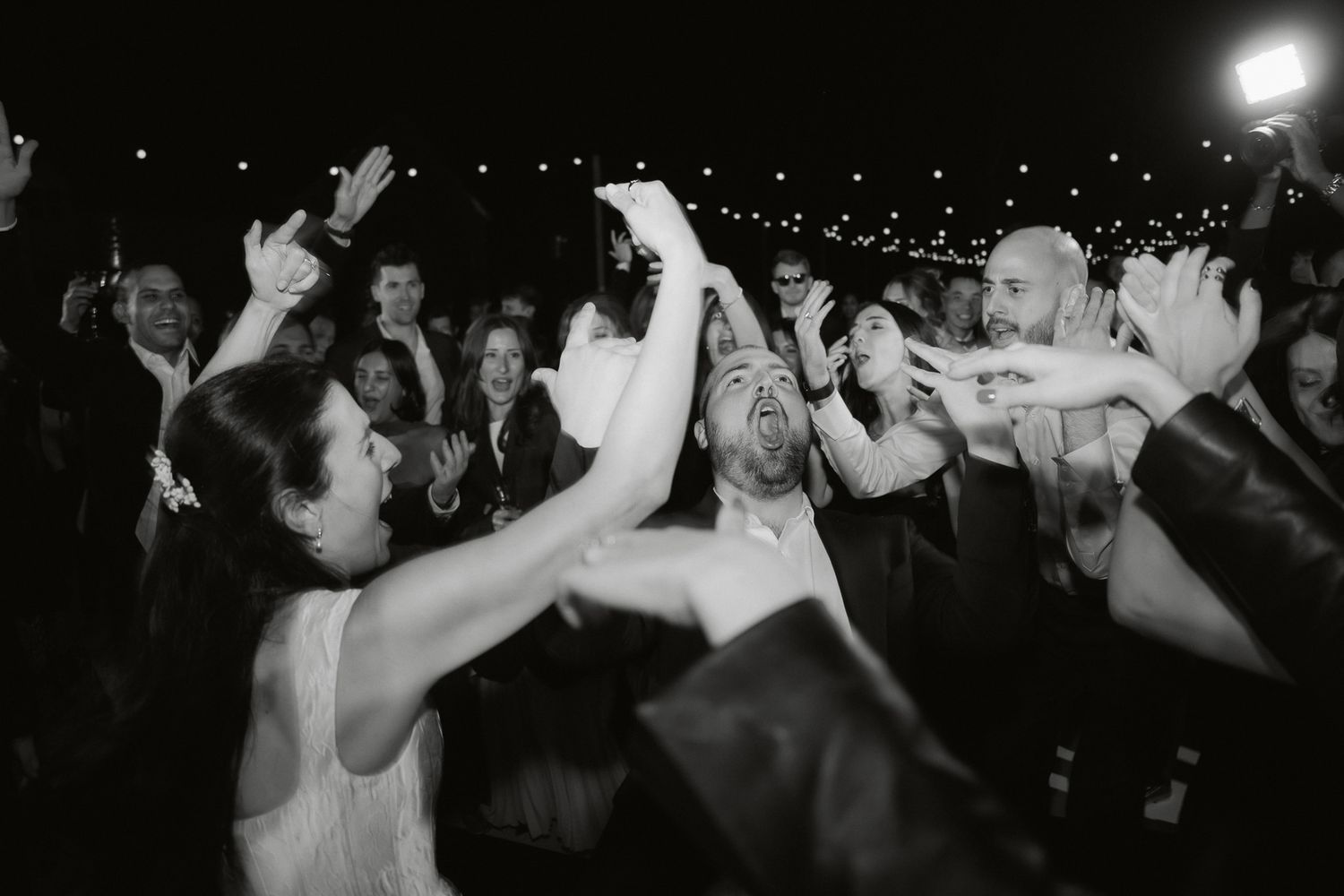 Black and white photo of joyful wedding guests dancing and celebrating at a lively outdoor reception at night.