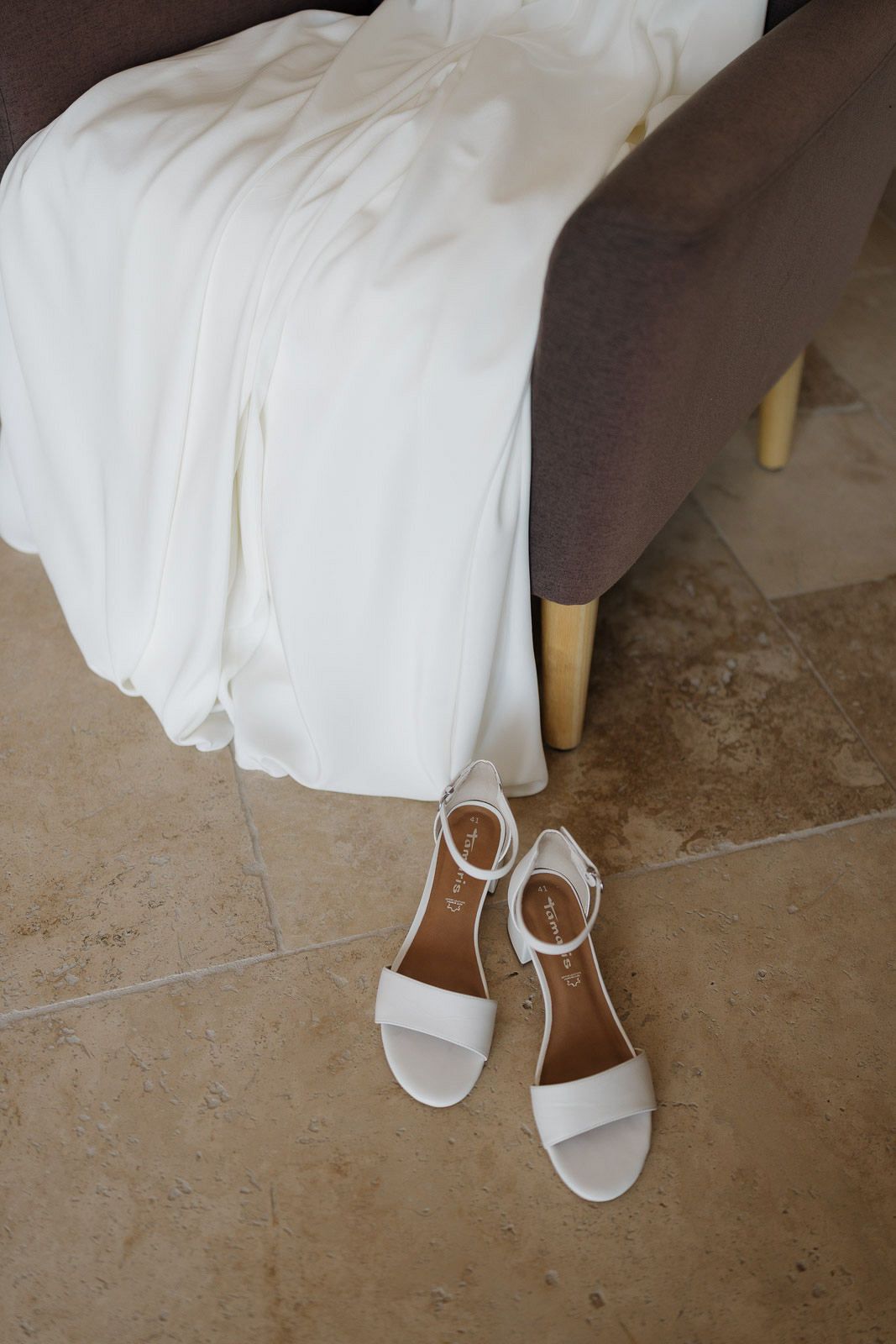 Close-up view of white bridal shoes peeking out from under a flowing wedding dress hem on a stone floor.