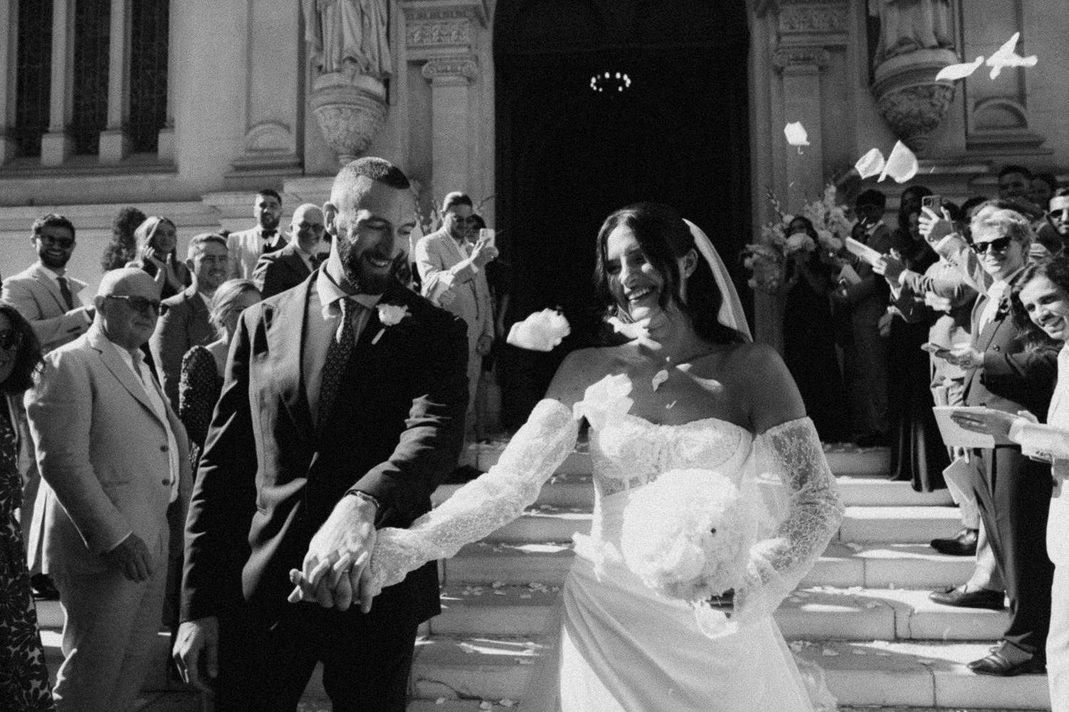A joyful bride and groom exit their wedding ceremony while guests shower them with confetti in black and white.