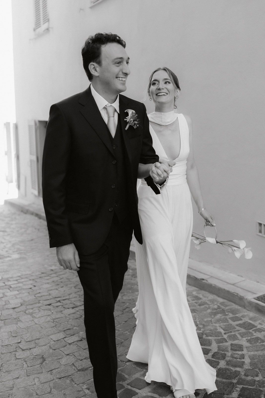 A black and white wedding photo of a bride in a sleek white gown and groom in a dark suit walking together on a cobblestone path.
