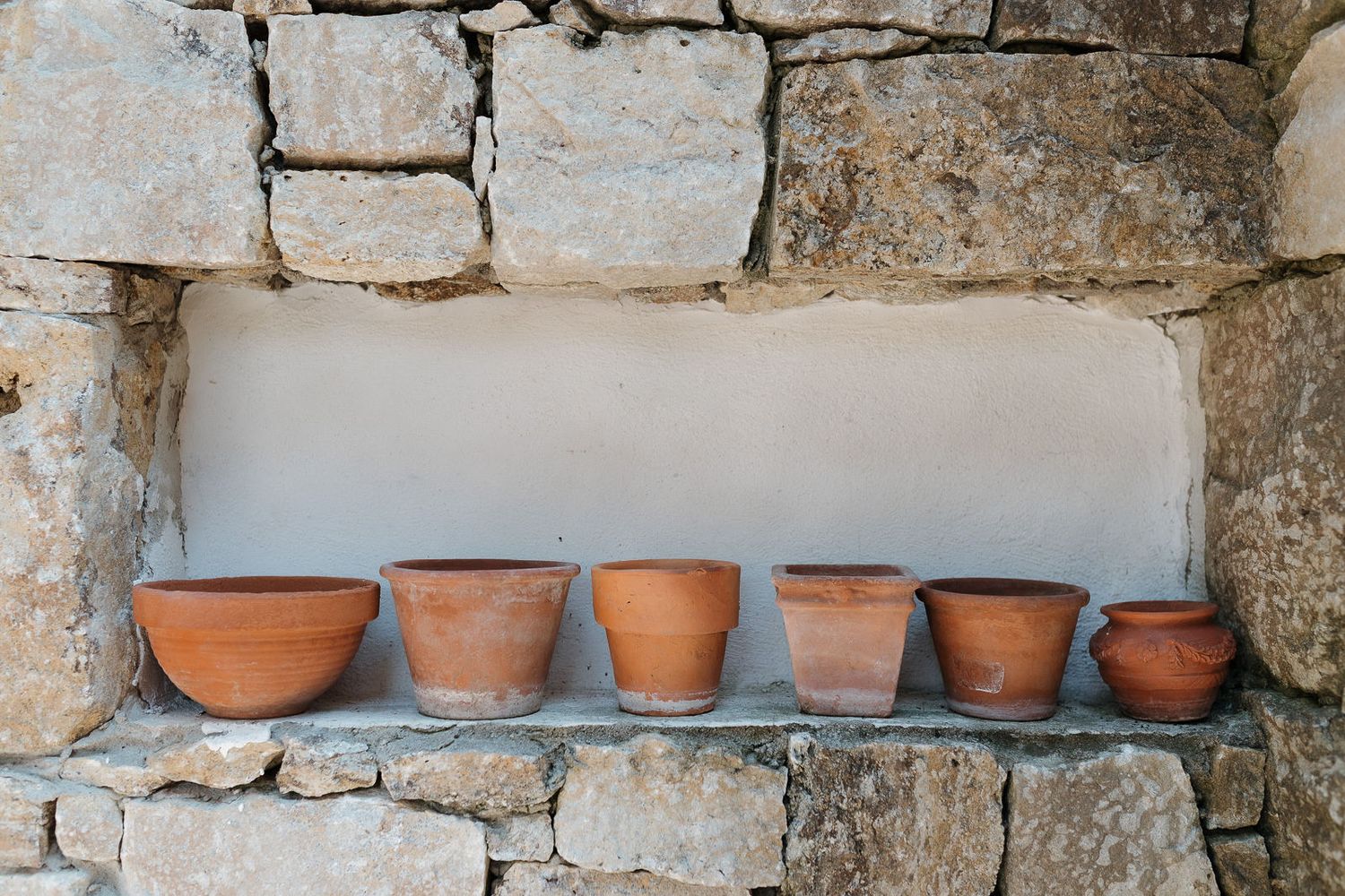 Rustic stone wall with small terra cotta pots arranged on a narrow shelf creating a Mediterranean garden display.