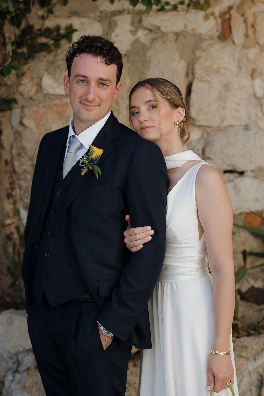 A wedding couple poses against a rustic stone wall backdrop in formal attire.