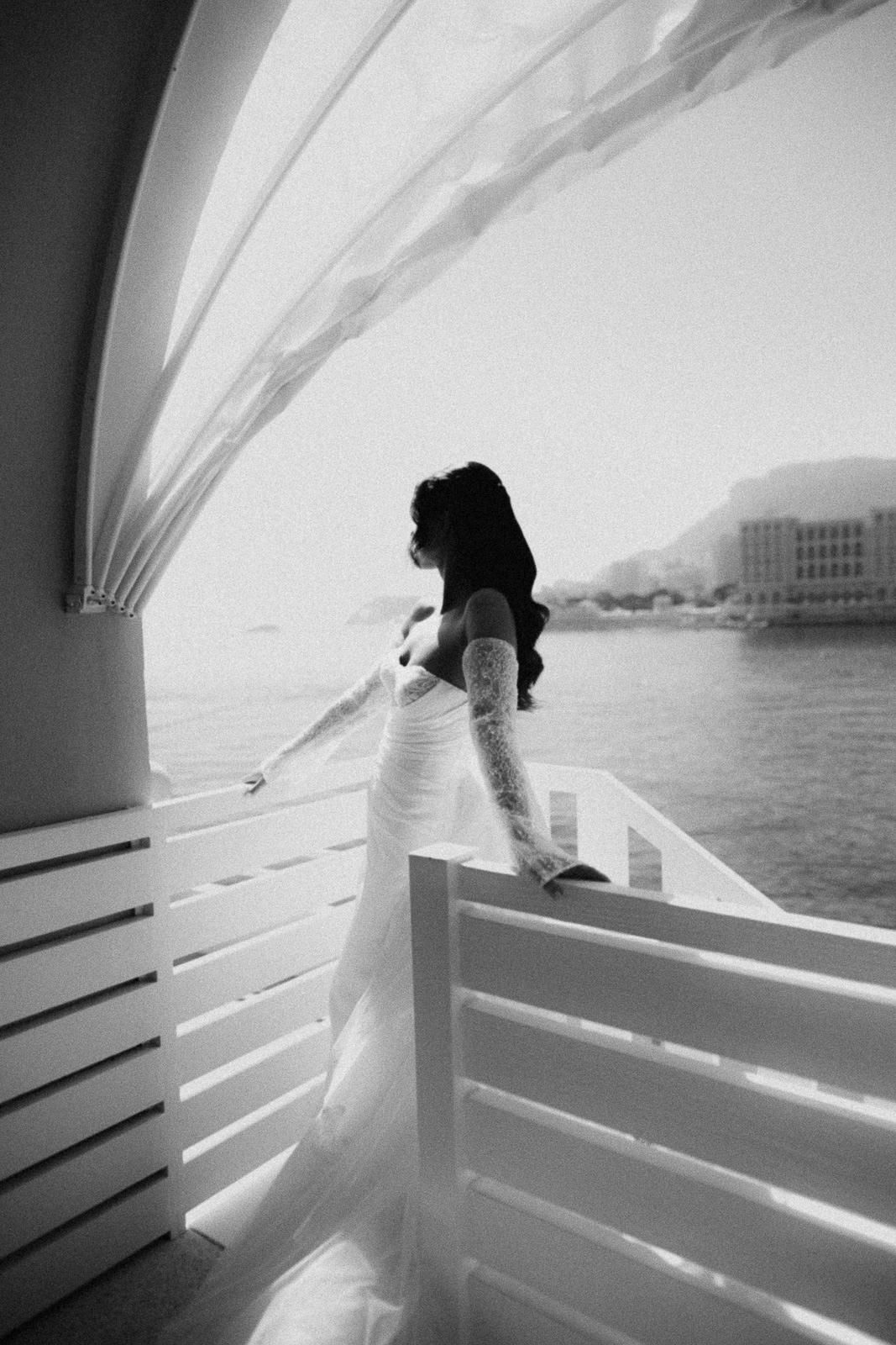 A silhouetted figure in a white dress stands on a boat deck overlooking the water on a bright day.