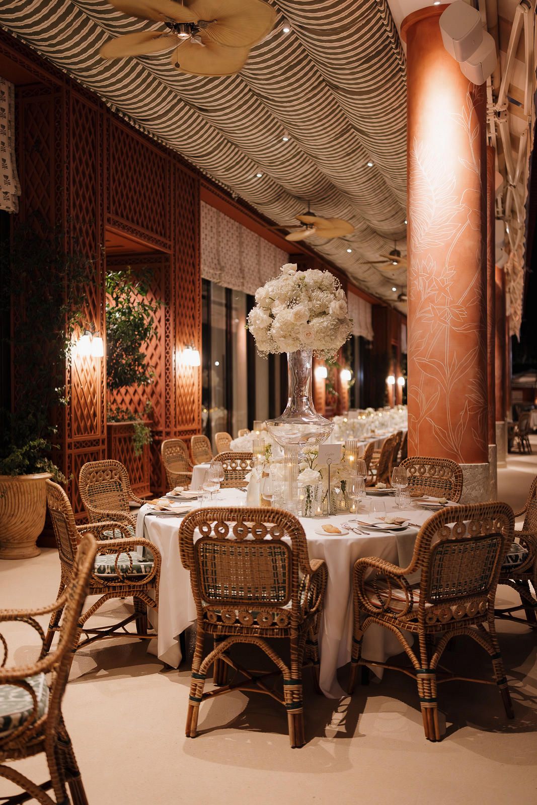 Luxurious restaurant interior with rattan chairs and white tablecloths under ornate ceiling with ambient lighting.