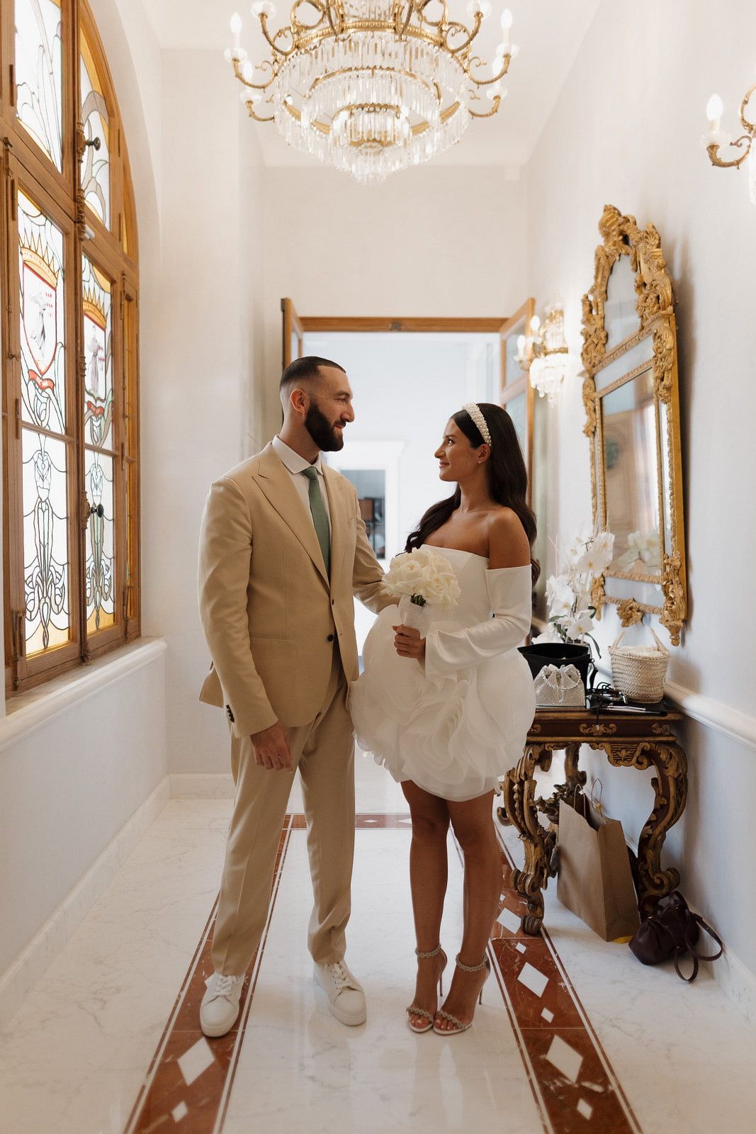 Couple in matching beige and white outfits pose together in an elegant white hallway with ornate gold mirrors.