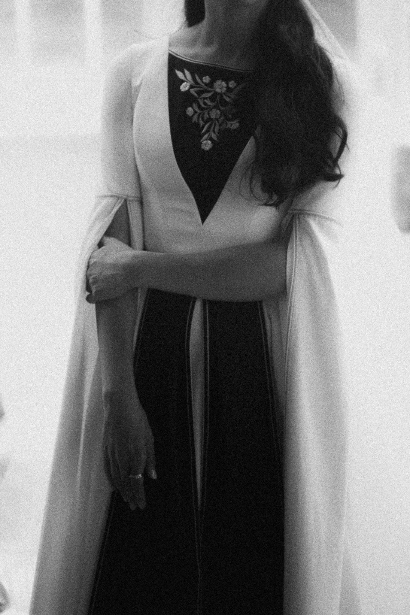 Elegant woman in a black and white floral embroidered gown with a flowing cape overlay.