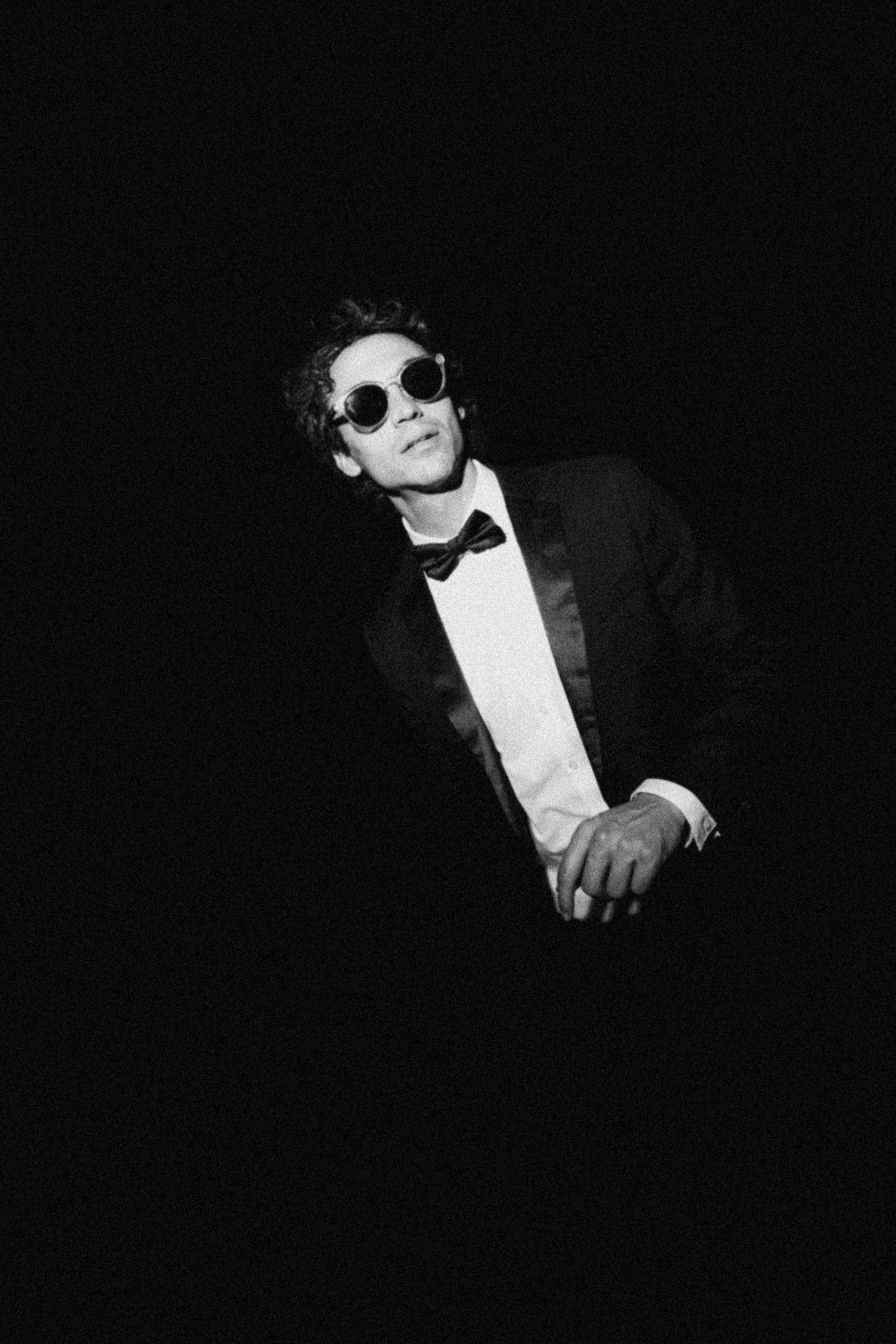 Black and white photo of a stylish man in a tuxedo and round sunglasses posing against a dark background.