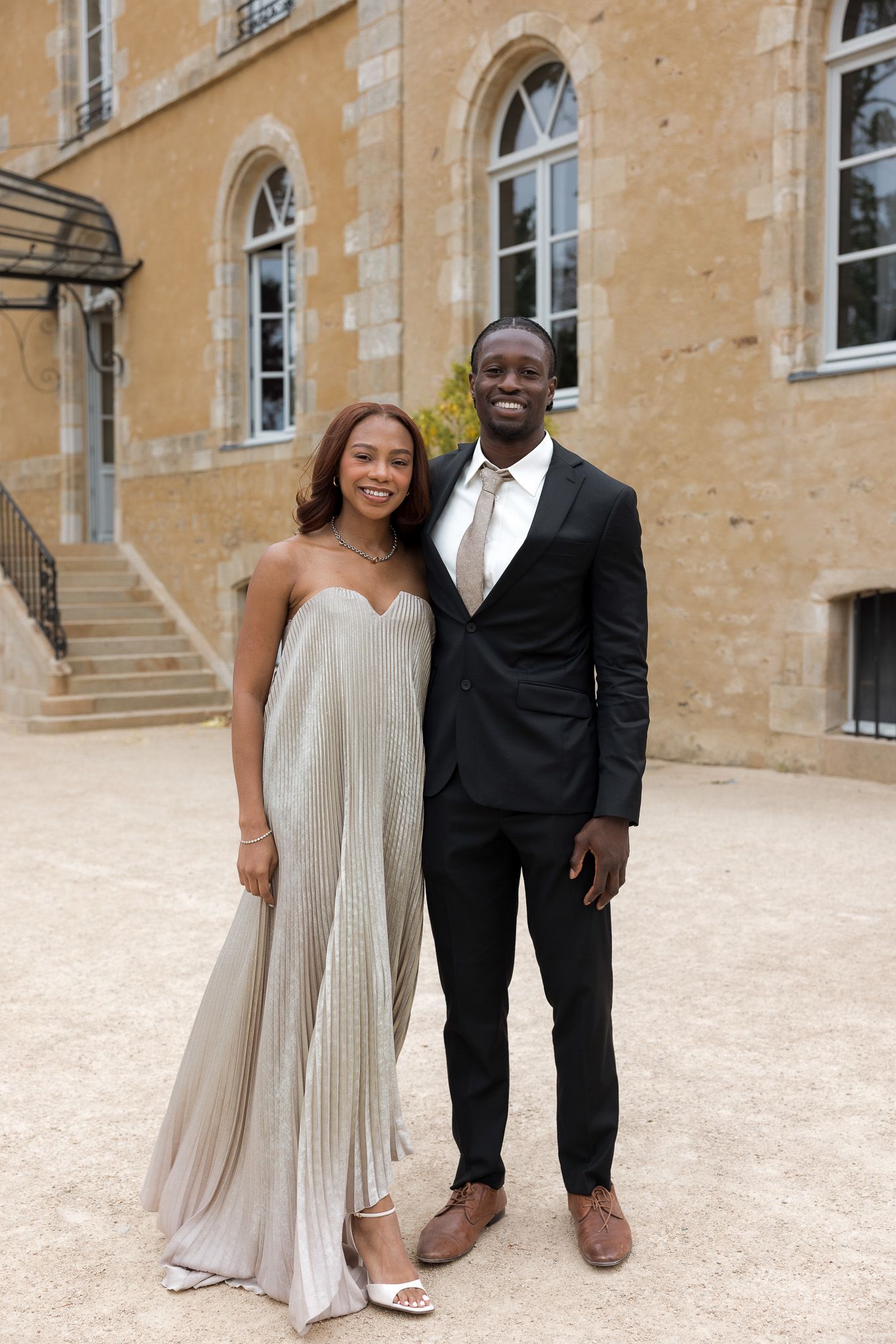 Elegant couple posing in formal attire outside a stone building, woman in silver gown, man in black suit.