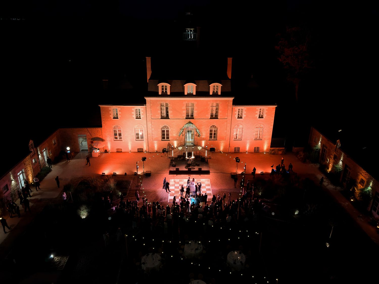 Aerial night view of an illuminated French chateau glowing orange, surrounded by crowds at an elegant outdoor event.