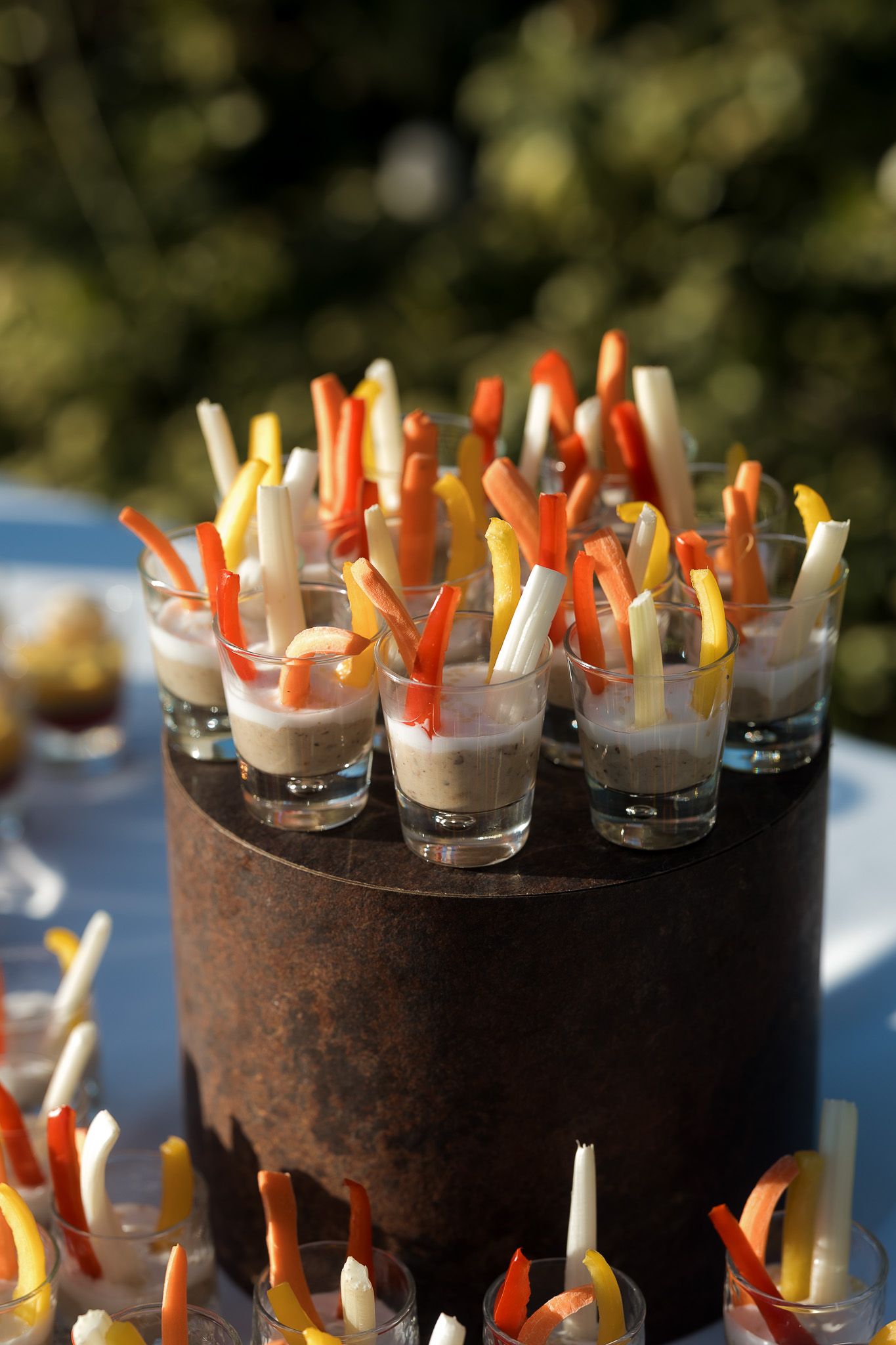 Elegant appetizer display with colorful vegetable sticks in shot glasses arranged on a dark cylindrical stand at an outdoor event.