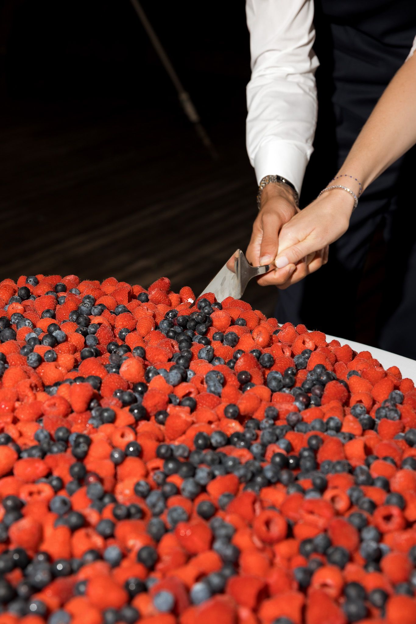 Two people cutting a large cake topped with fresh raspberries and blueberries using a knife.