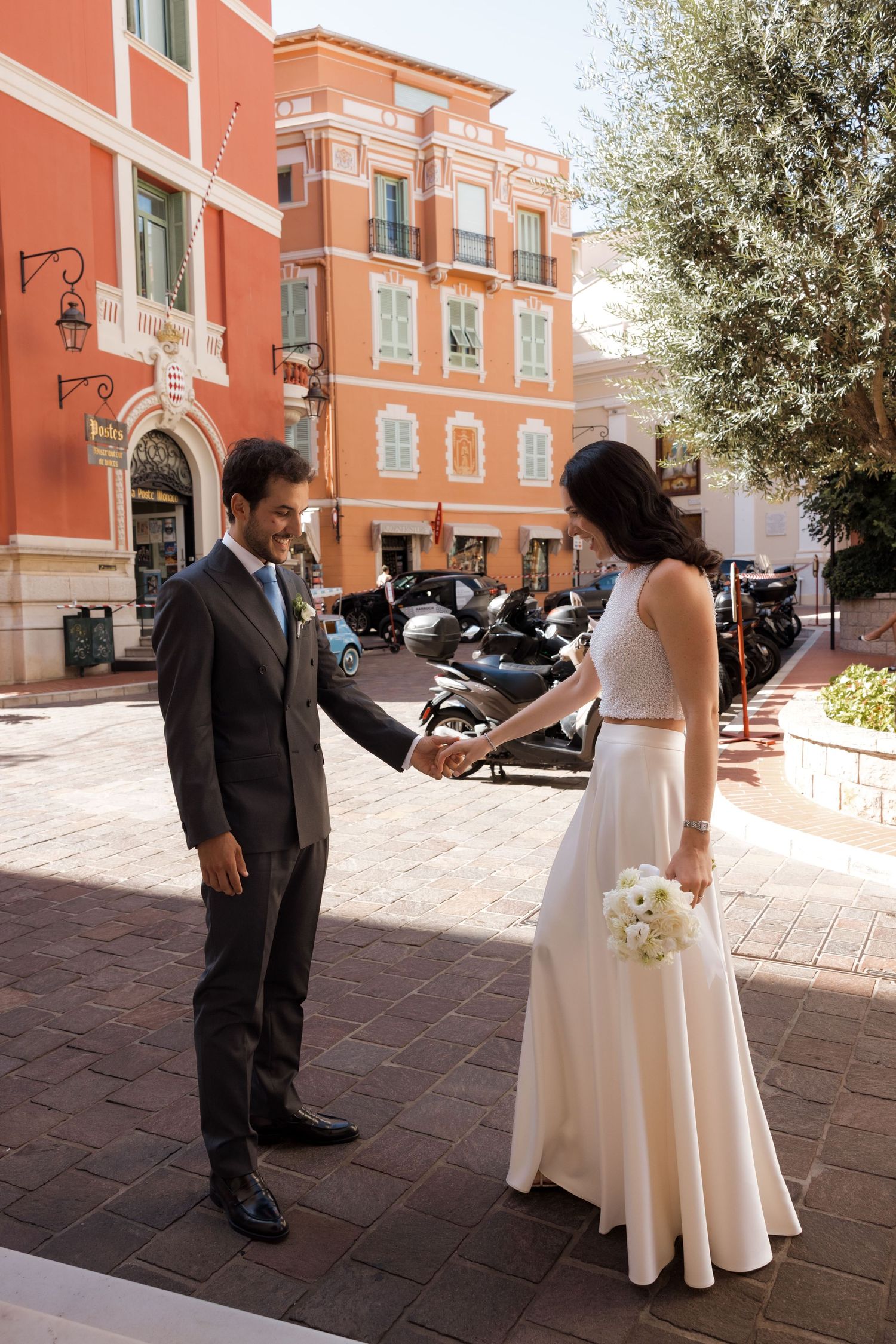Bride and groom hold hands on a sunny European cobblestone street, bride holding white bouquet in elegant two-piece wedding gown.