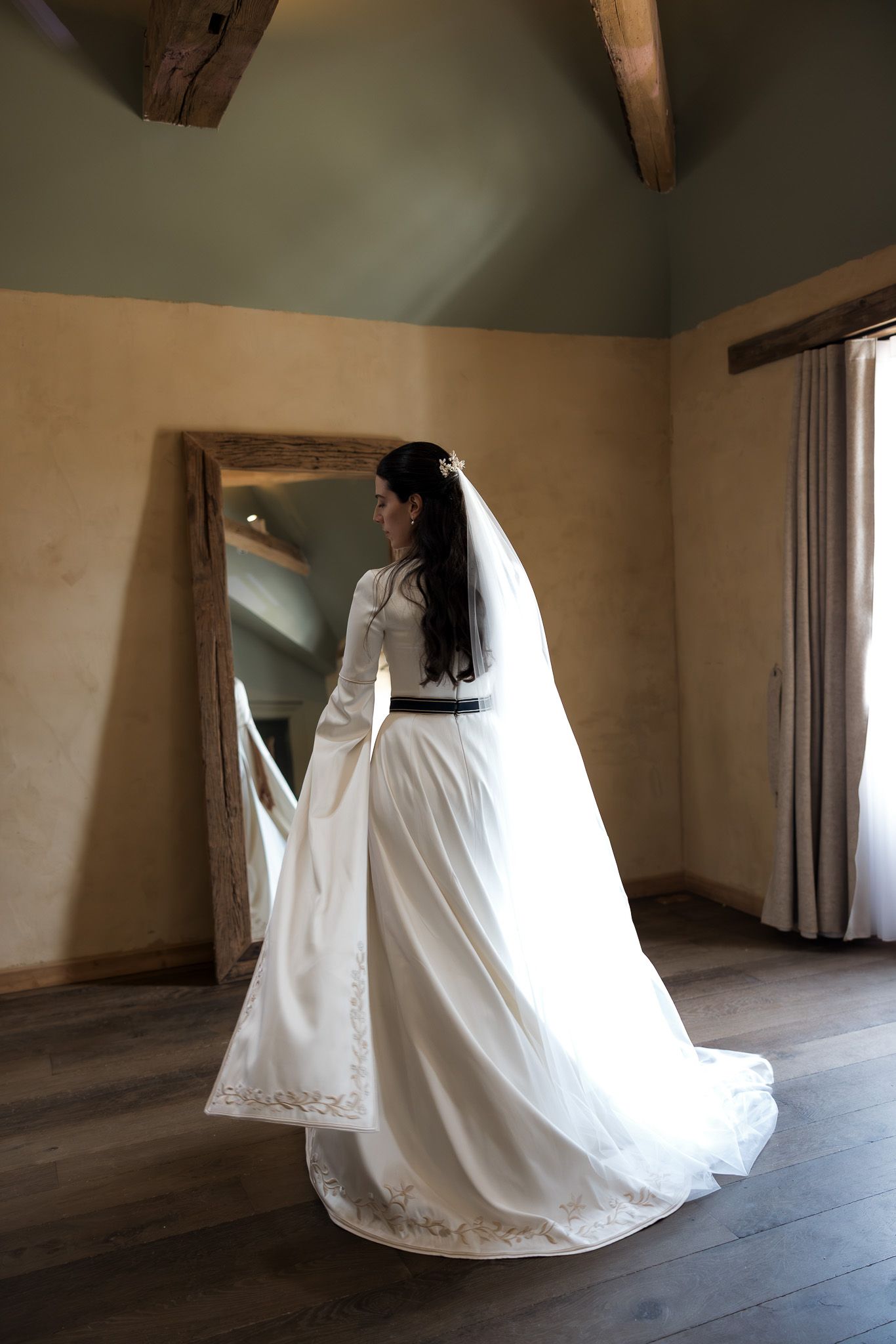Bride in elegant white wedding gown with long veil and black belt stands before a large mirror in a rustic room.