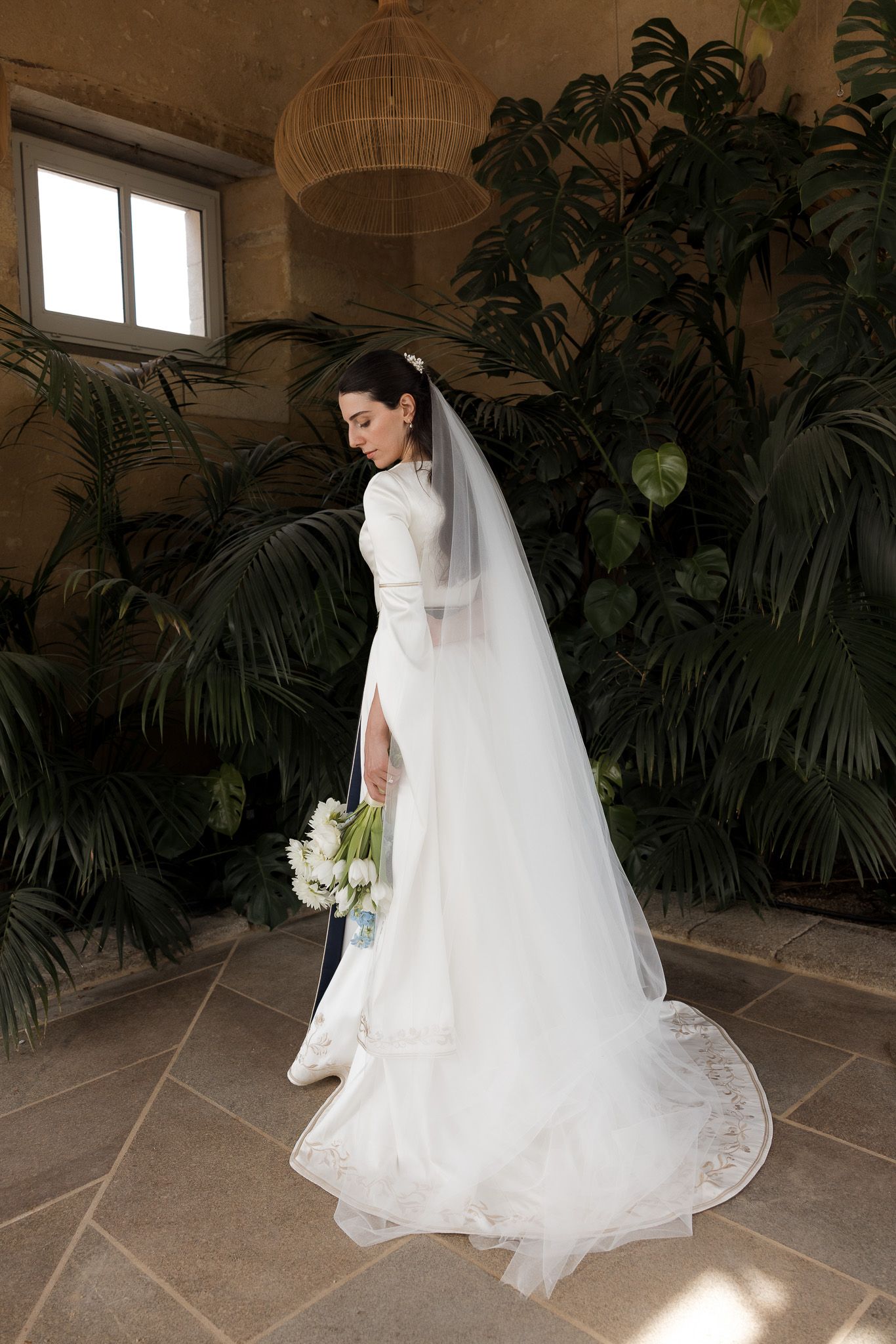 Elegant bride in a white long-sleeve wedding gown with veil holding a bouquet, standing among lush tropical plants indoors.